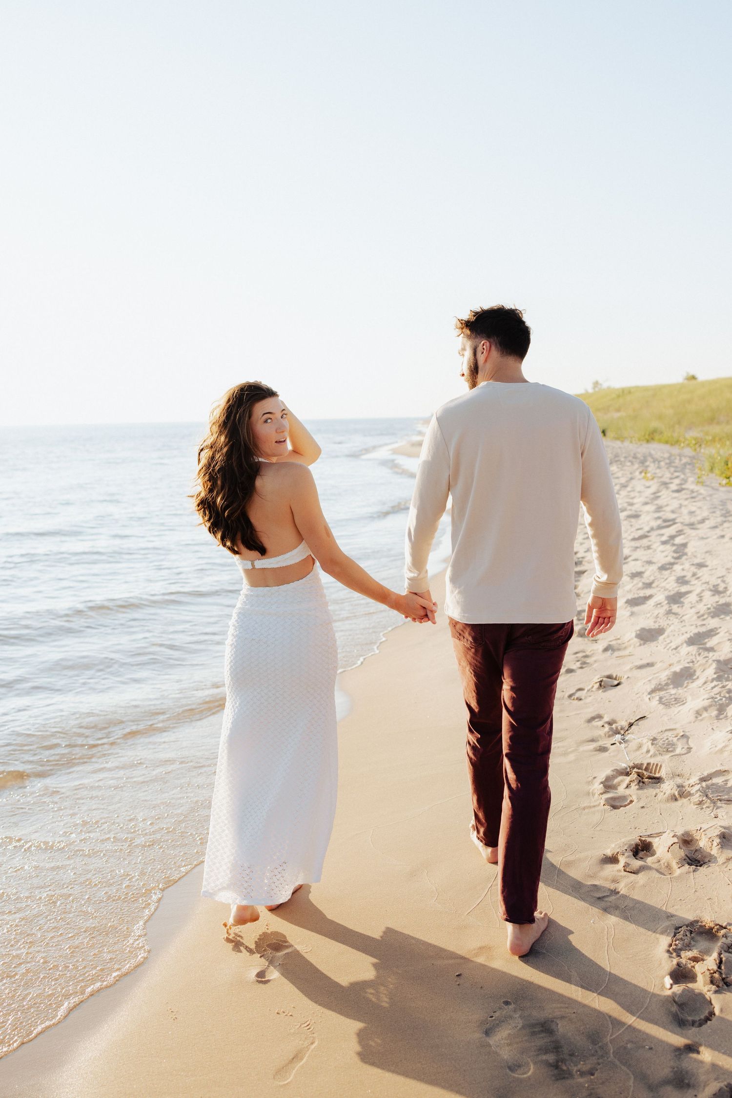 Couple holding hands walking barefoot on sandy beach, woman in white dress looking back, sunny day.