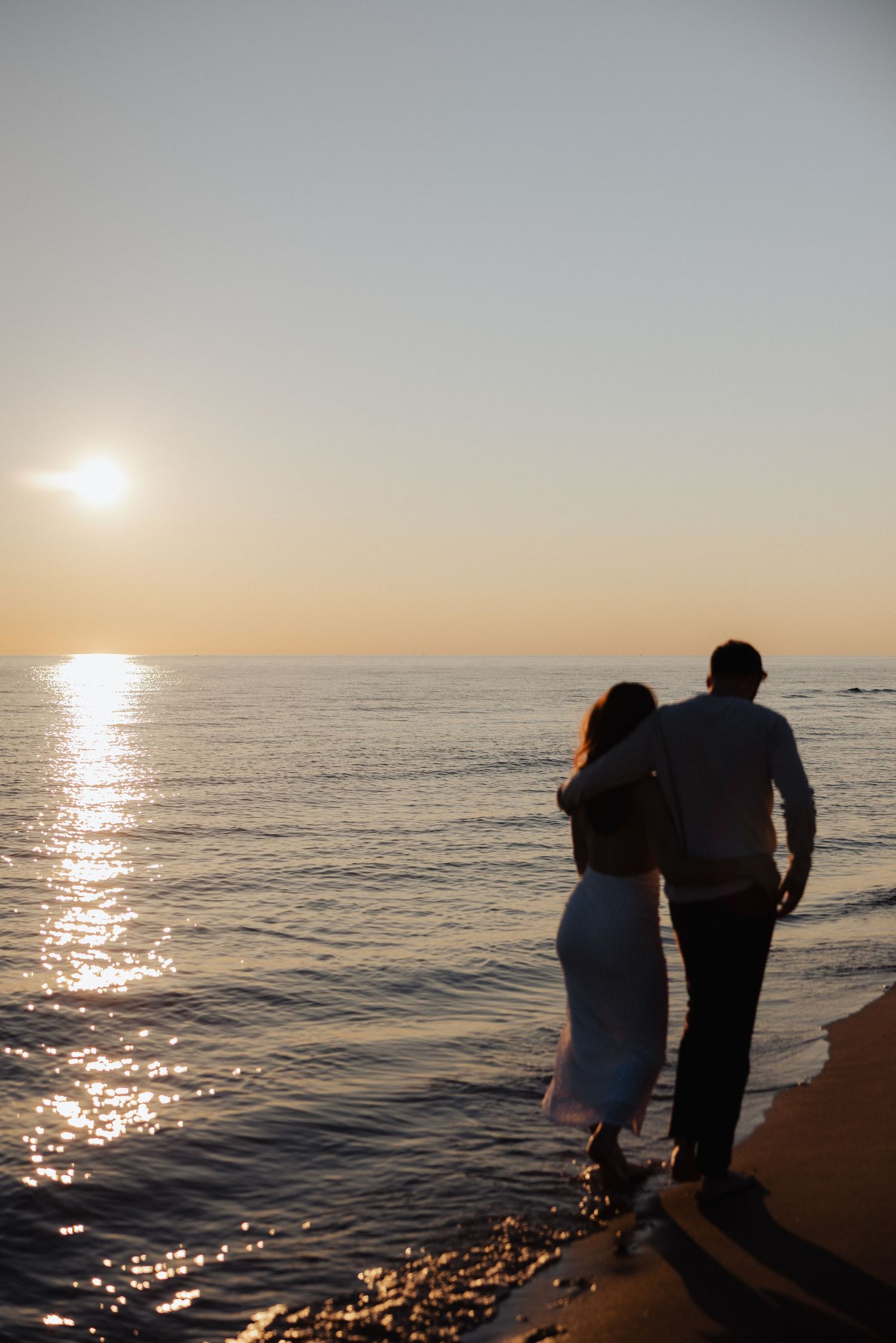 Couple embracing on sandy beach at sunset, silhouetted against golden ocean reflections and clear sky.