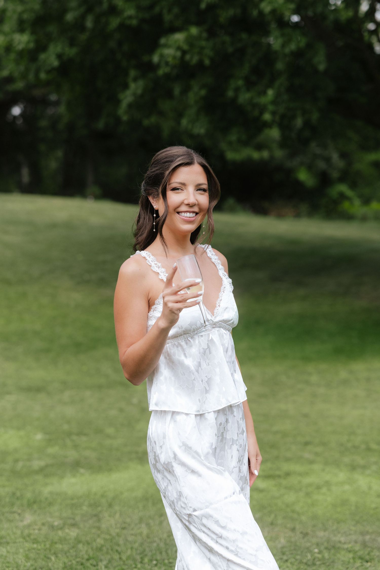 Smiling bride in white lace gown holding champagne glass outdoors on green lawn.