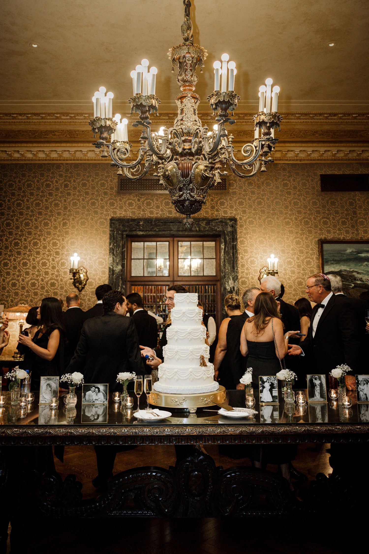 Guests in formal black attire surround a white tiered wedding cake beneath an ornate chandelier.