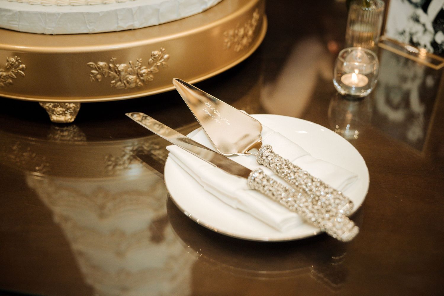 Elegant wedding cake serving set with crystal-encrusted handles on a white plate beside a gold cake stand.