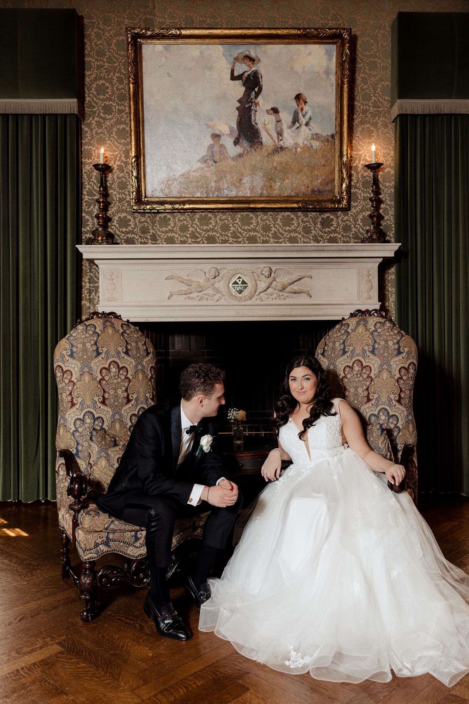 Bride and groom sit in ornate chairs by a fireplace beneath a classic oil painting in an elegant room.