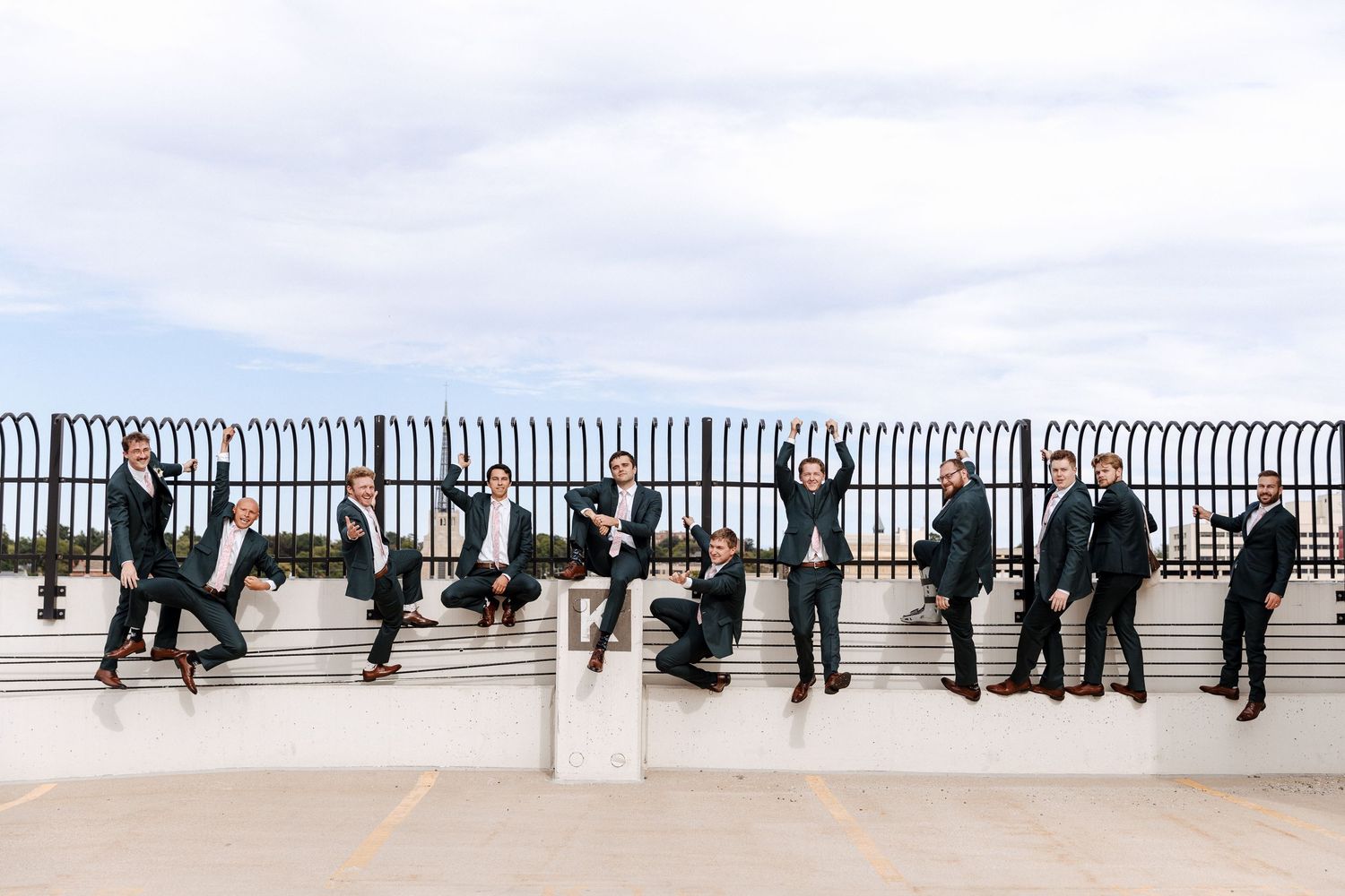 Groomsmen in black suits jump and pose playfully along rooftop railing under cloudy sky.