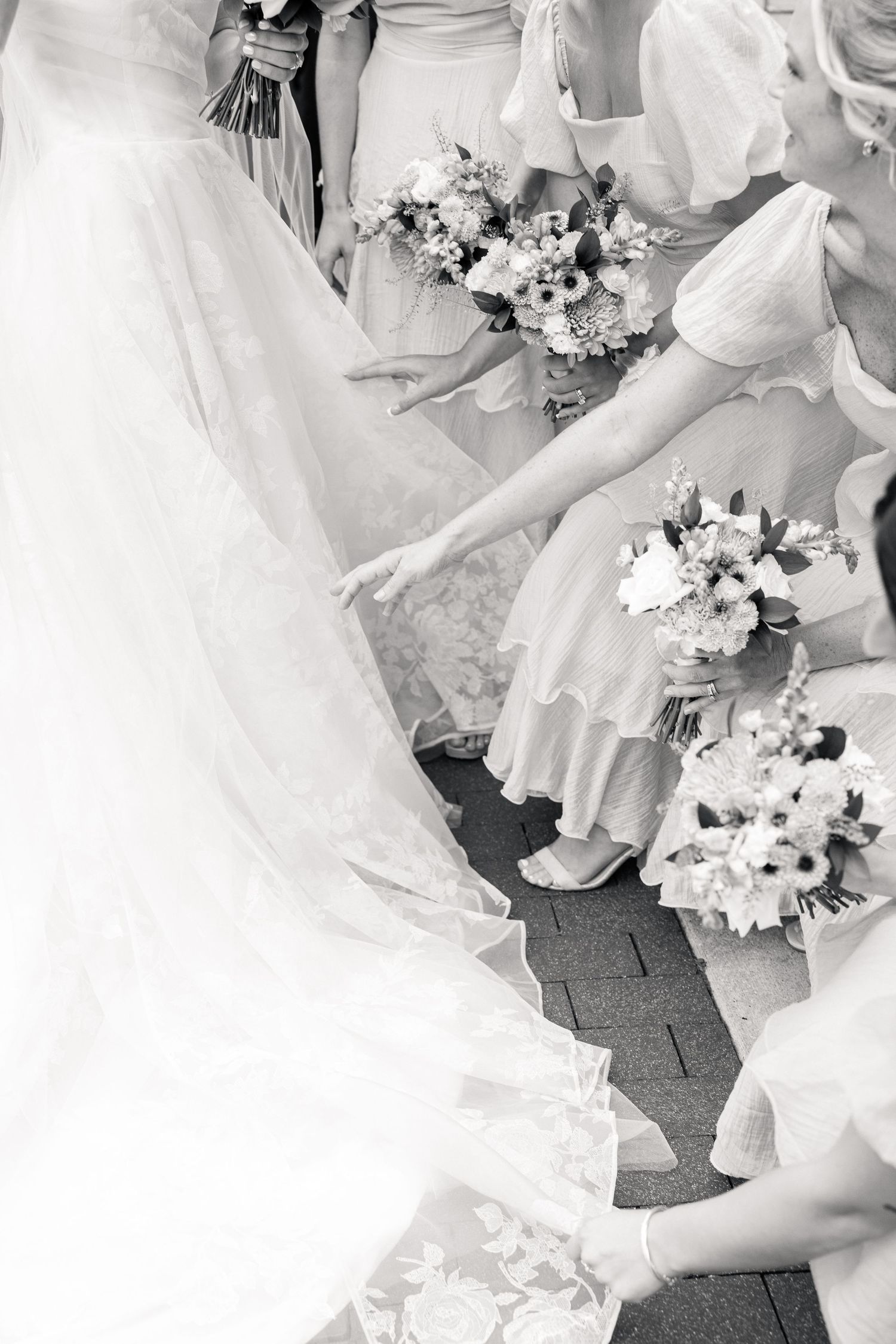Black and white photo of bridesmaids in ruffled dresses holding bouquets while arranging the bride's lace wedding train.