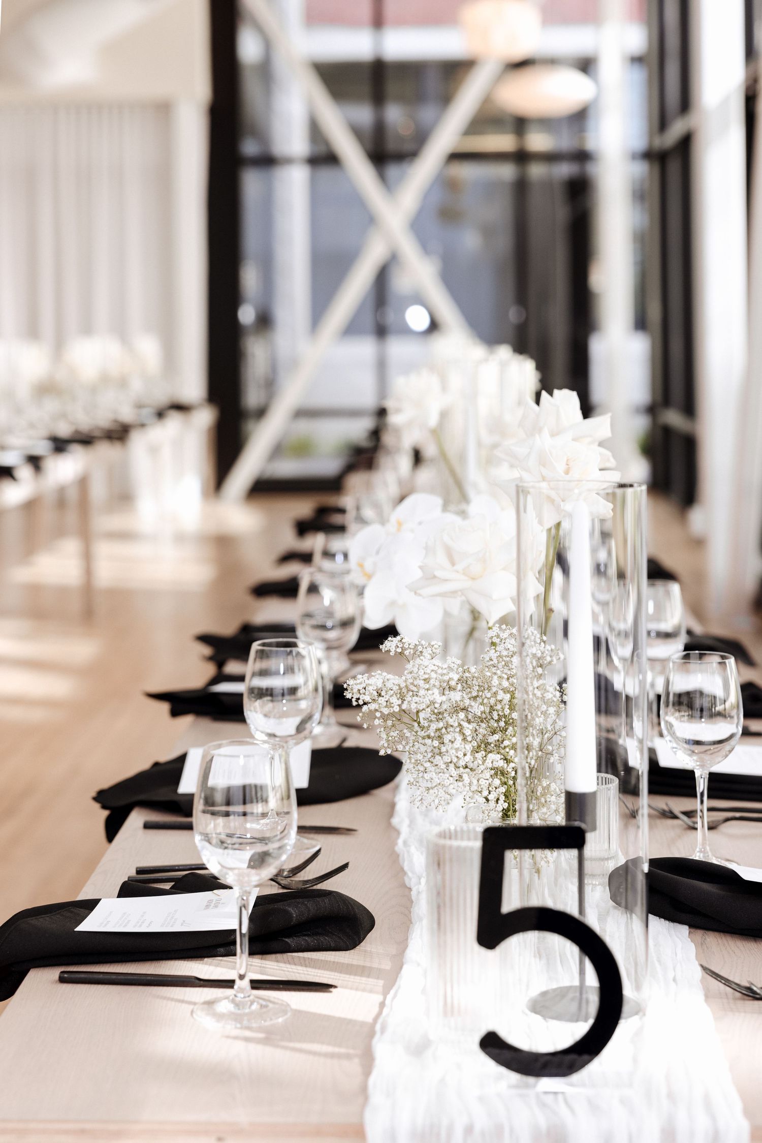 Modern wedding reception hall with long tables set with black napkins, white florals, and a bold number 5 table marker.