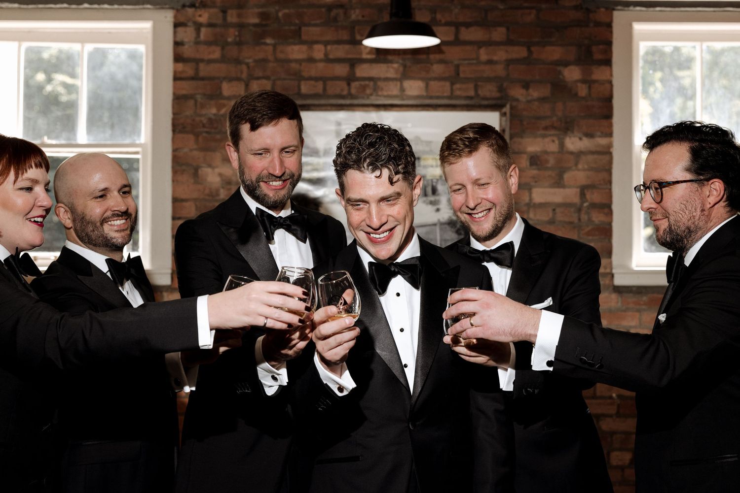 Groomsmen in black tuxedos toasting with whiskey glasses at a wedding celebration in a brick-walled venue.