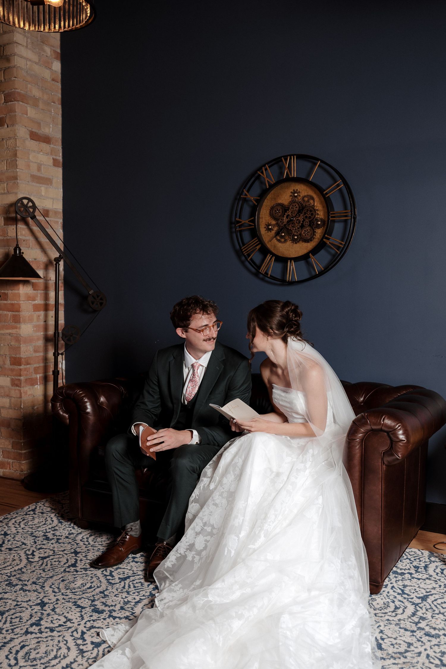 Bride and groom sit on leather sofa reading together beneath decorative clock on dark blue wall.