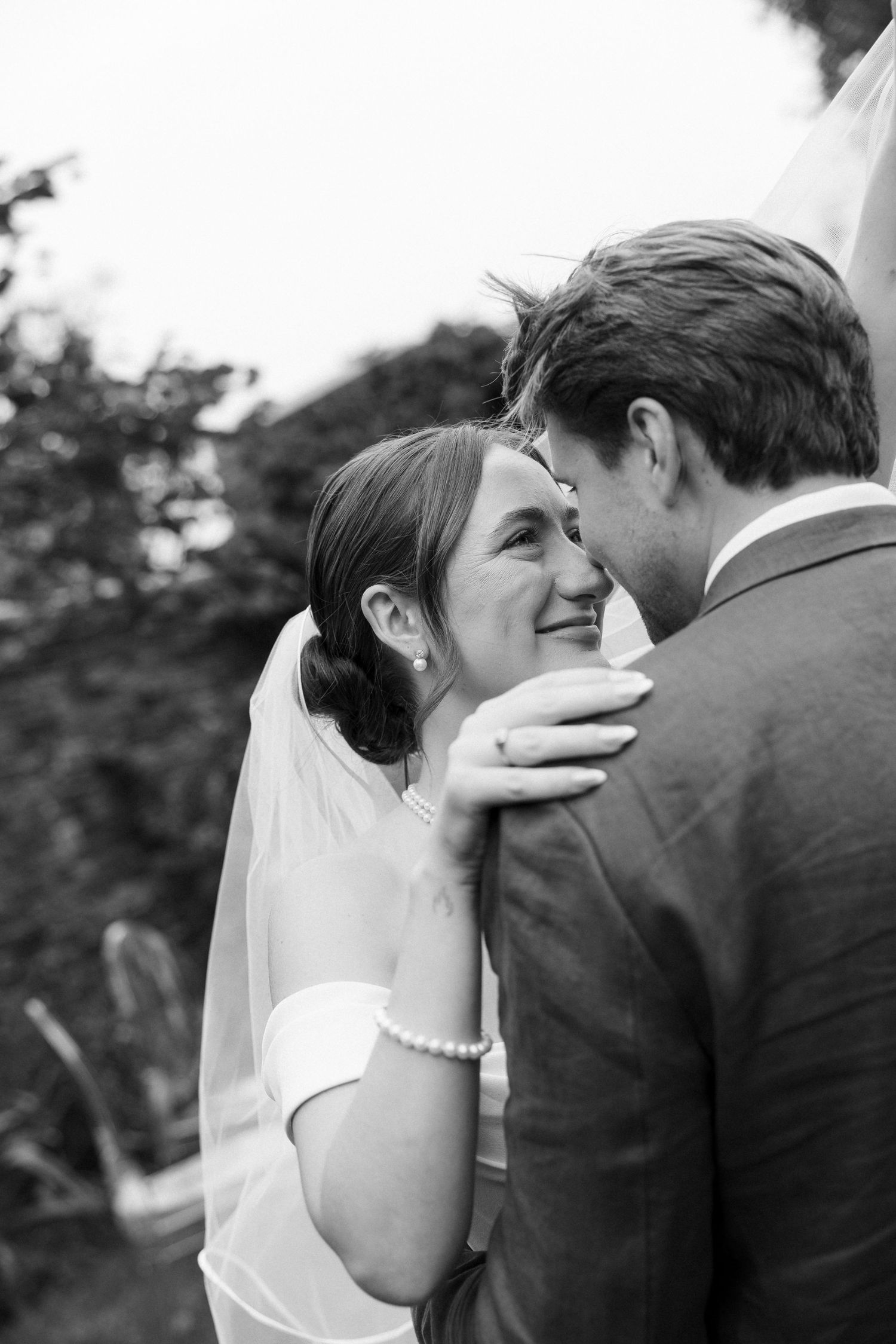 Black and white wedding photo of smiling bride in veil and groom sharing an intimate moment outdoors.