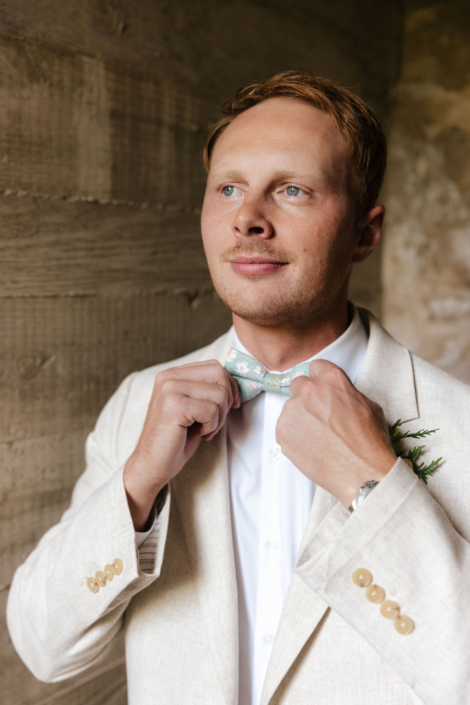 Groom in ivory linen suit adjusting light blue polka dot bow tie with boutonniere at wedding.