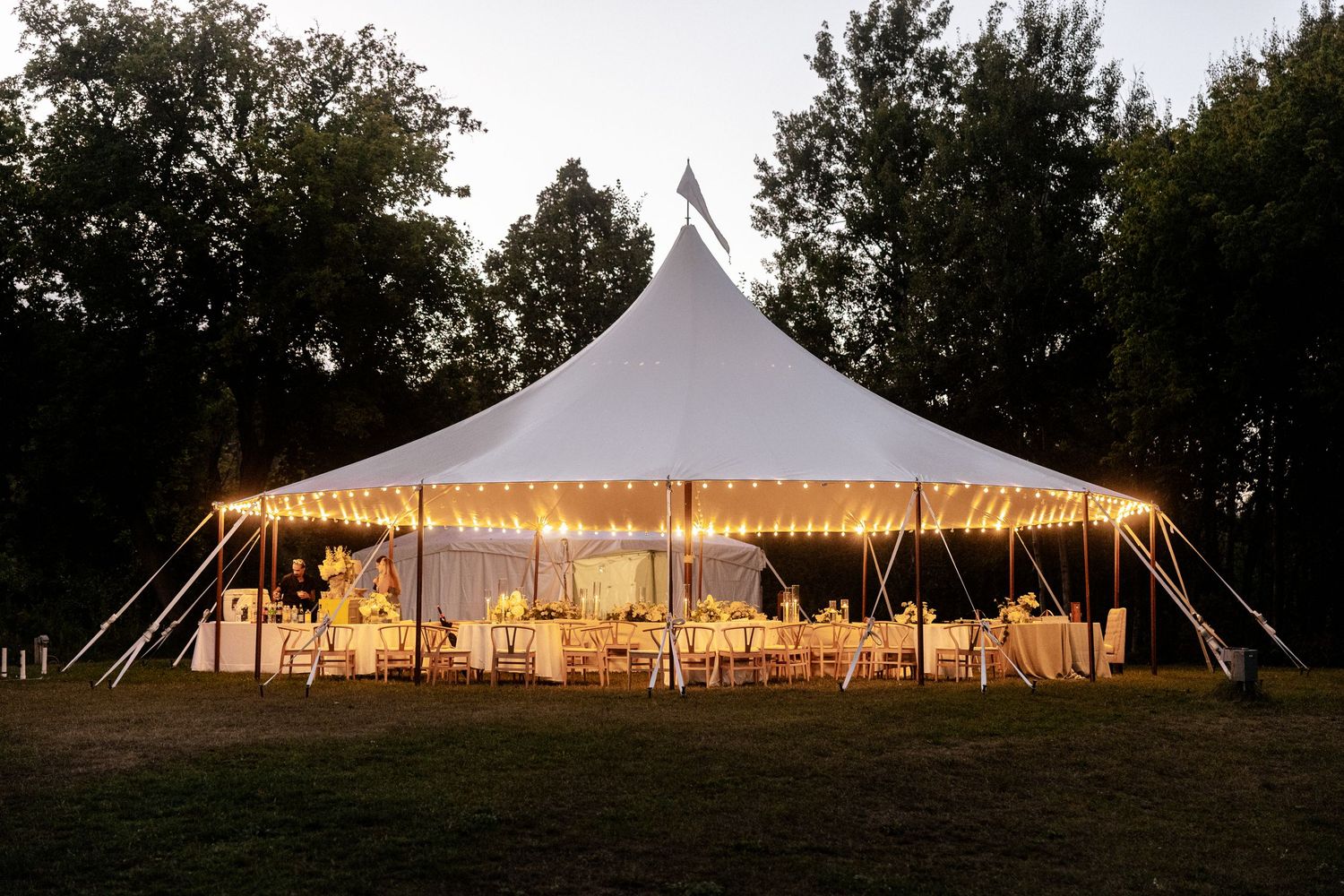 Elegant white sailcloth wedding reception tent glowing with warm lights at dusk surrounded by tall trees.