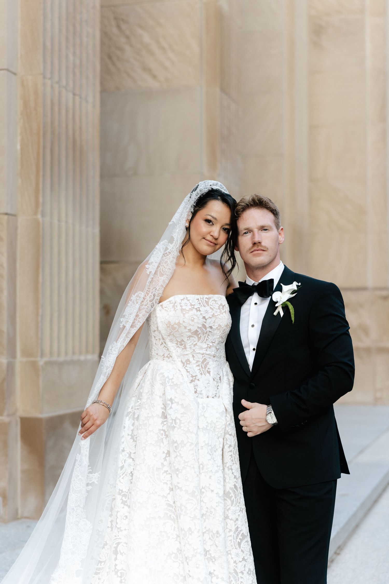 Bride in lace gown and veil poses with groom in black tuxedo before stone columns at elegant wedding venue.