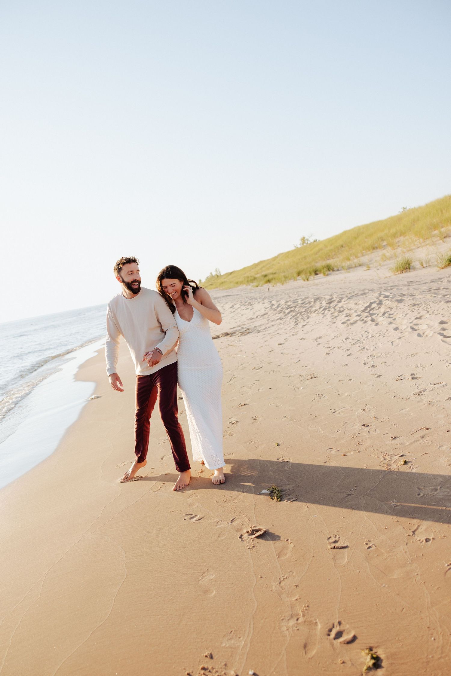 A couple walks hand-in-hand along a sandy beach, woman in white dress, man in dark pants, with dunes and clear sky behind.