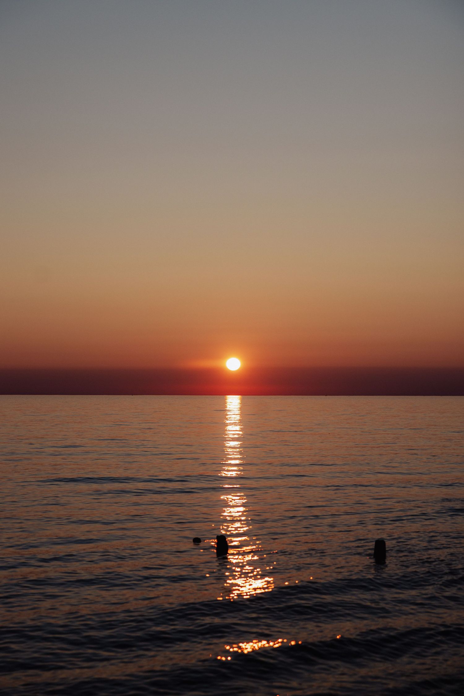 Stunning ocean sunset with golden reflection on calm water and silhouettes of swimmers along the horizon.