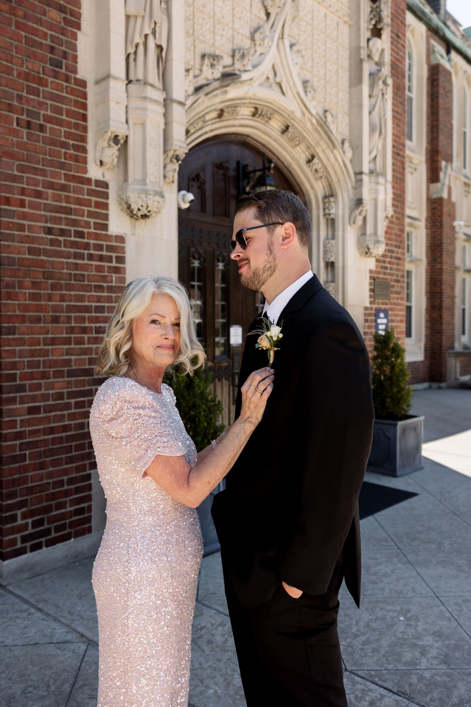 Elegant mother adjusting groom's boutonniere in black tuxedo outside brick church on wedding day.