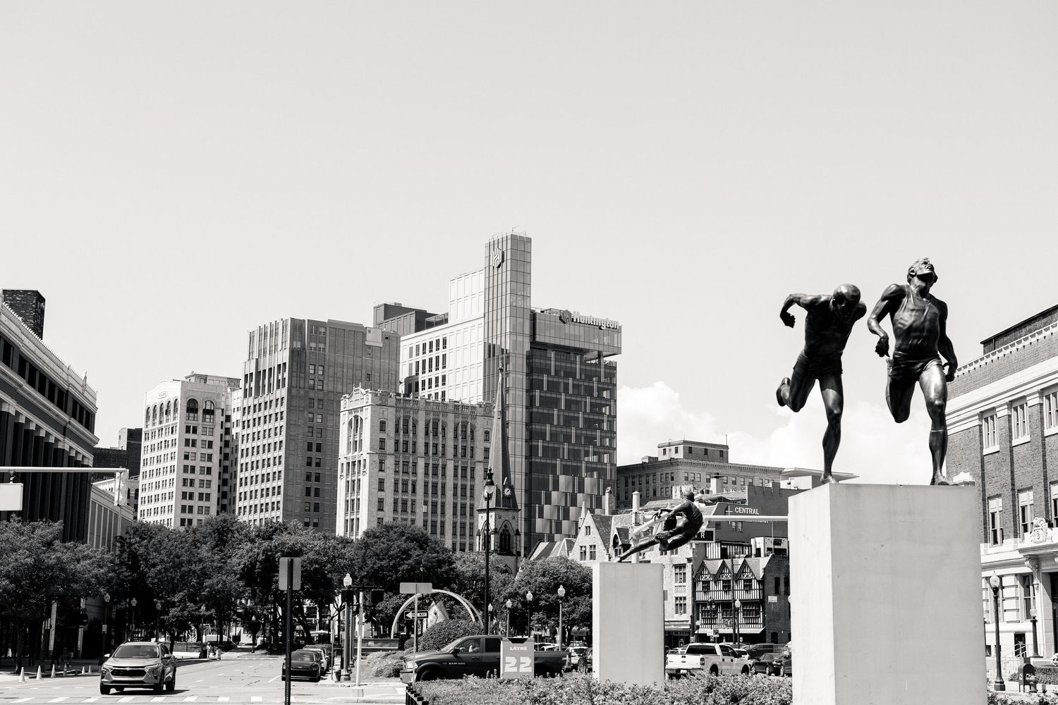 Black and white cityscape with bronze statues on pedestals along a busy urban street.