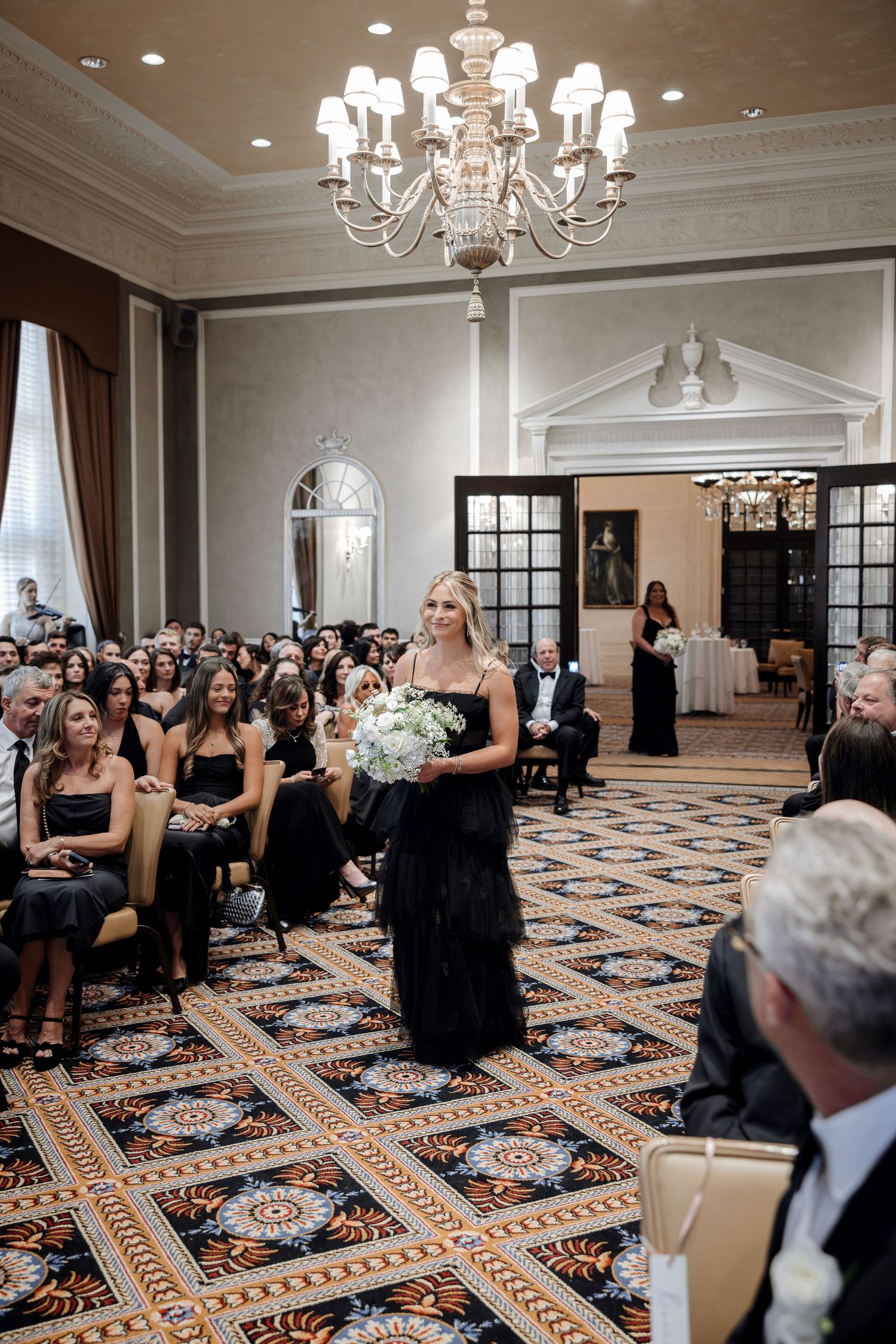 Bridesmaid in black dress walks down the aisle carrying white bouquet in an elegant ballroom wedding ceremony.
