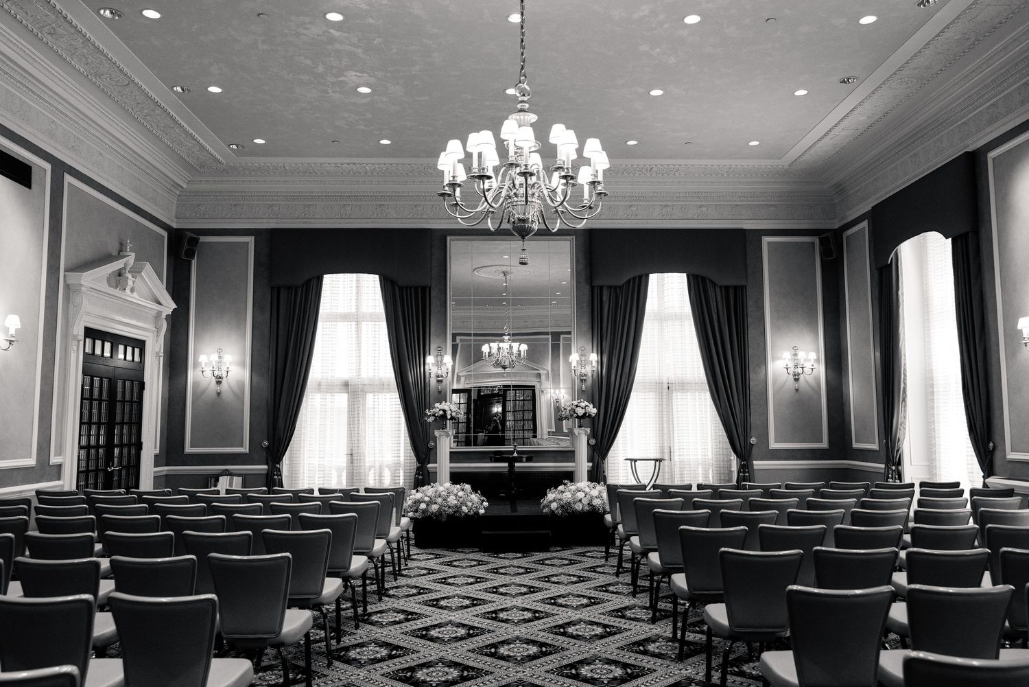 Black and white image of a grand ballroom wedding ceremony venue with chandelier, tall windows, and rows of chairs.