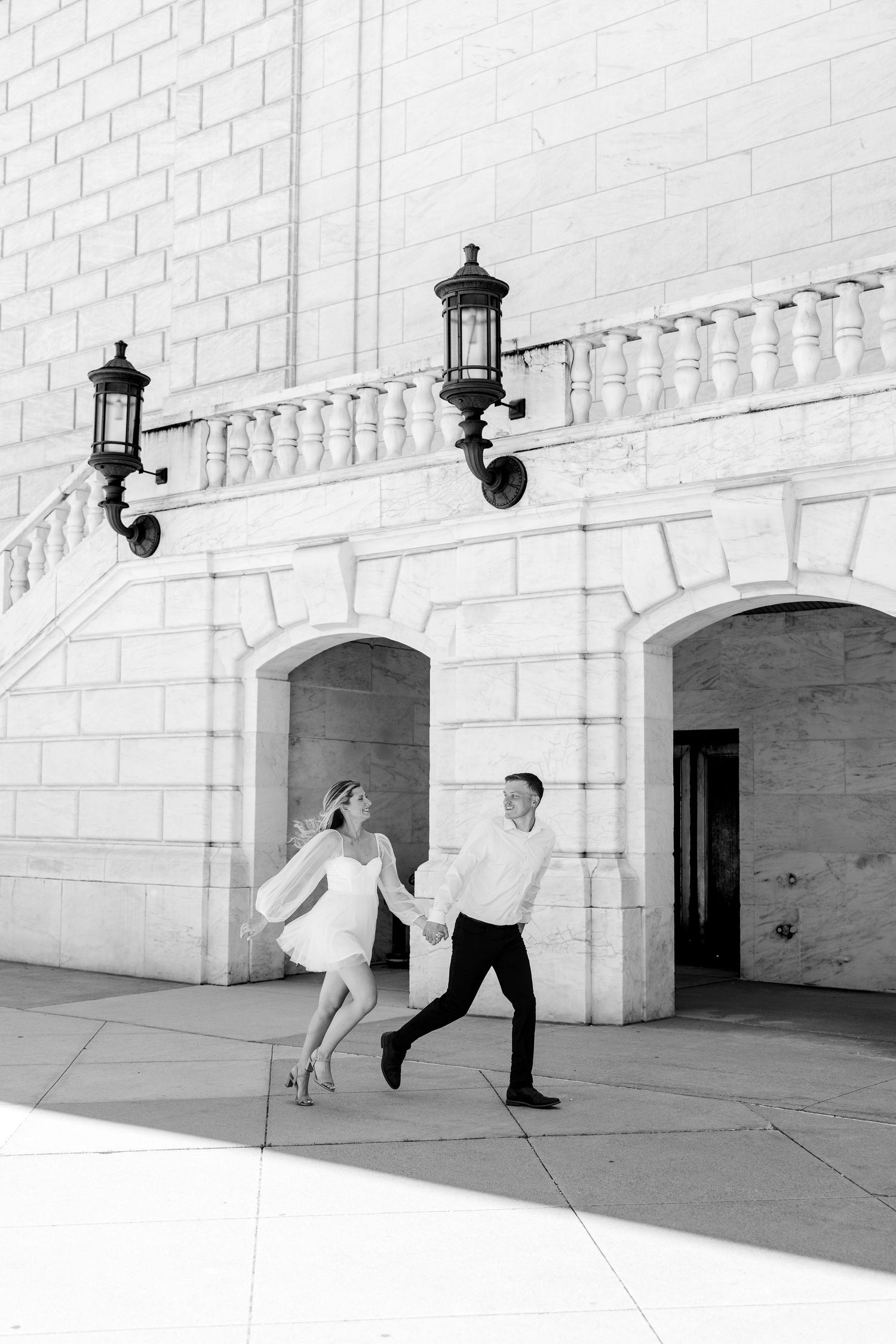 Black and white photo of a couple holding hands and running joyfully in front of a grand white stone building with arched doorways.