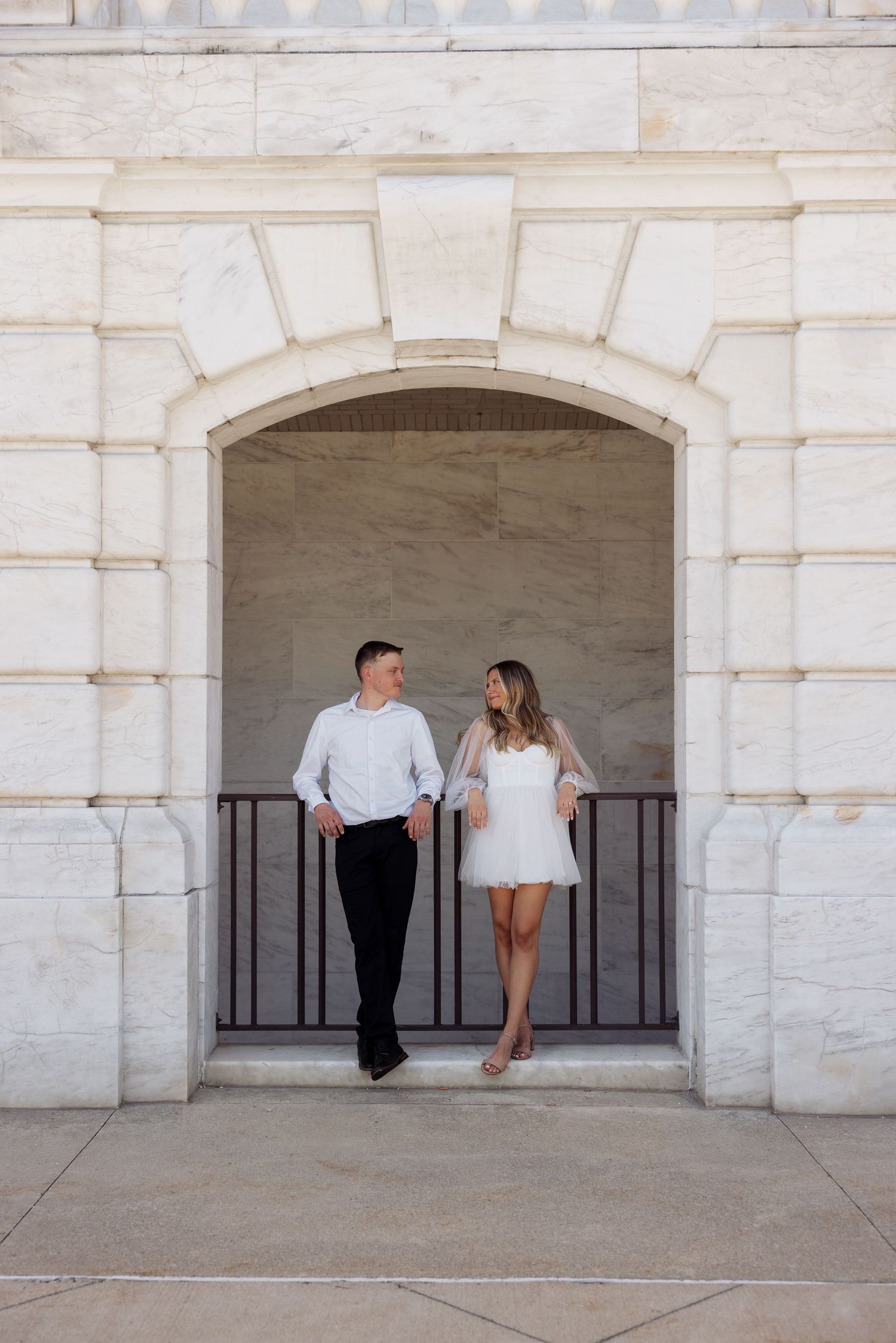 Couple in white outfits standing together under a stone archway at an elegant marble building.