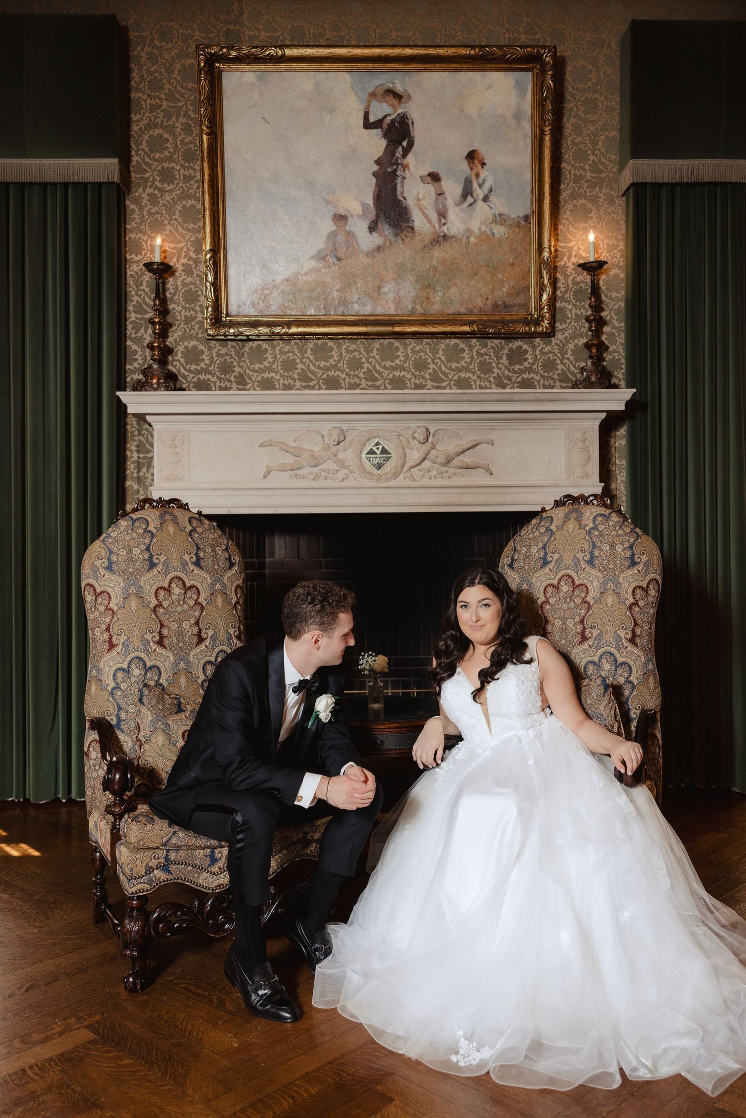 Bride and groom sit in ornate chairs by a fireplace, holding hands, with a classic painting and candles behind them.