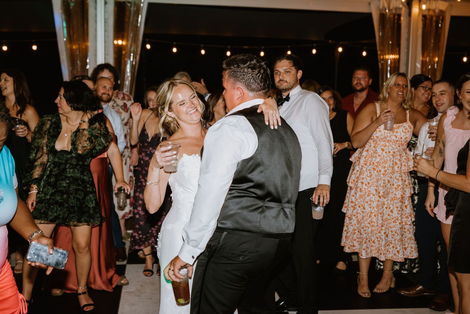 Bride and groom share a joyful dance surrounded by guests at an outdoor wedding reception with string lights.