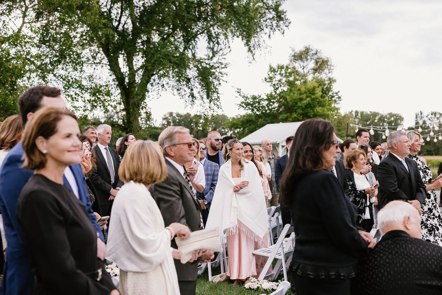Guests gathered outdoors for an elegant wedding ceremony on a lush green lawn beneath a large shade tree.