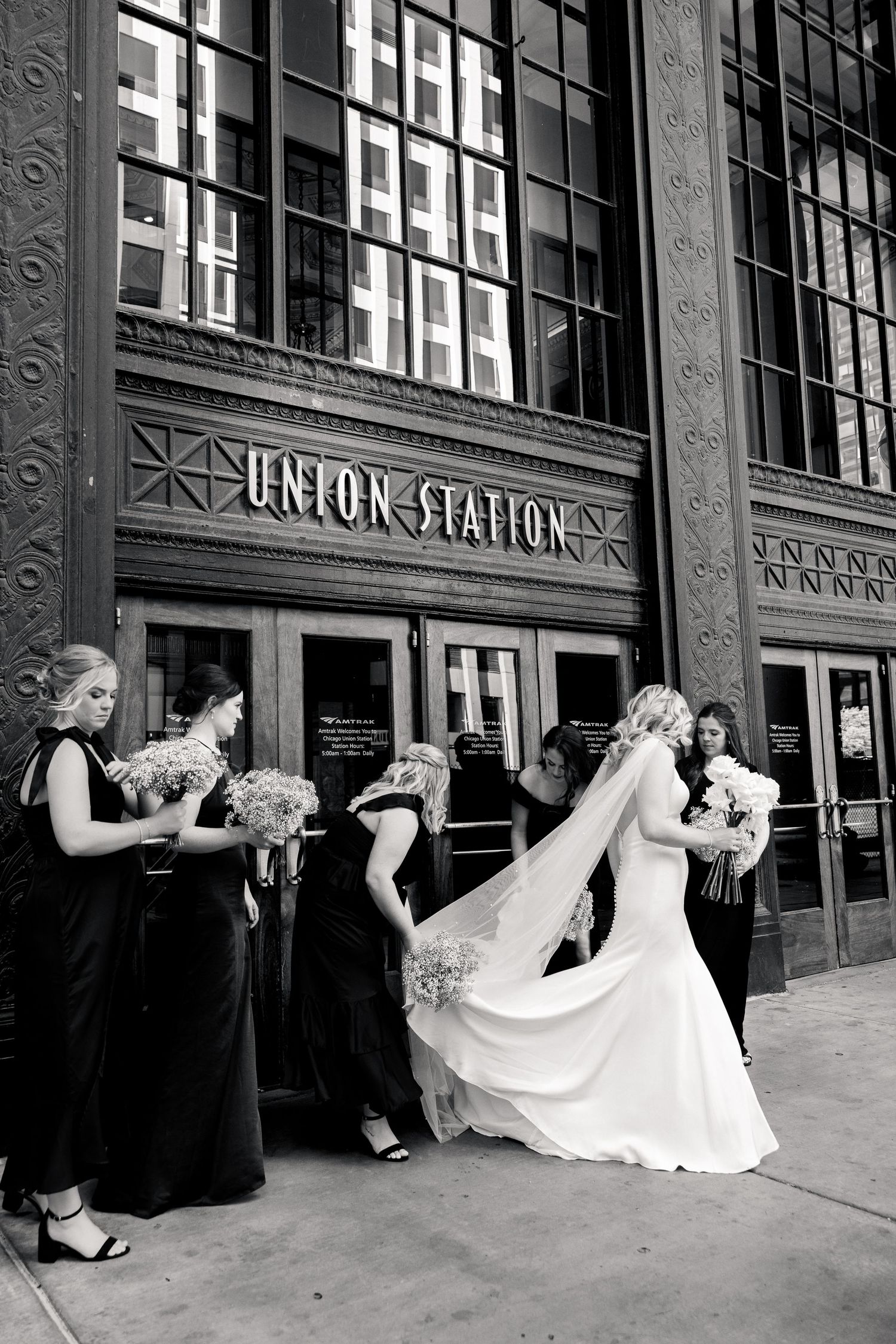 Bride and bridesmaids in black dresses pose outside Union Station in a classic black and white wedding photo.