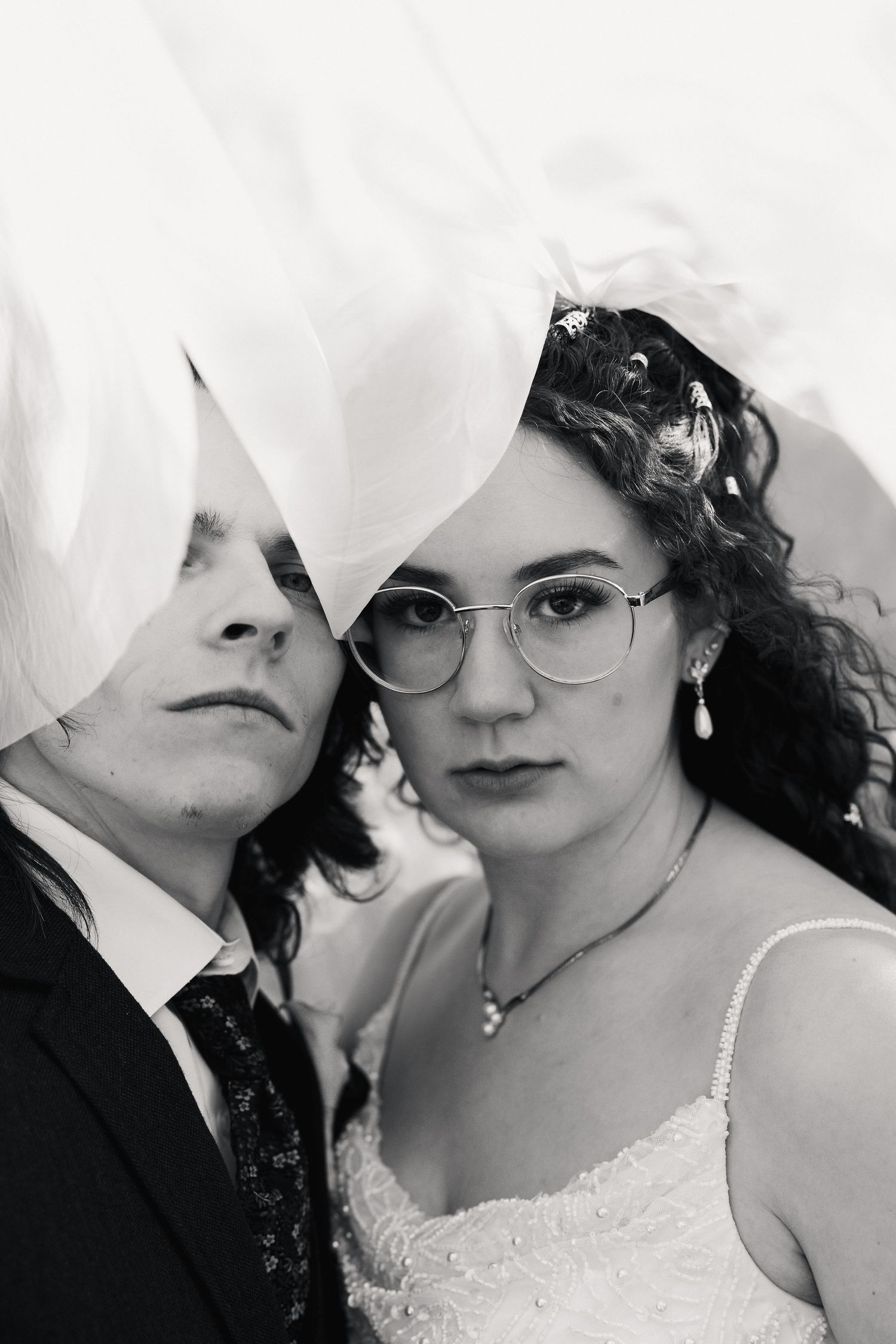 Black and white wedding portrait of bride in glasses and groom partially covered by flowing veil.