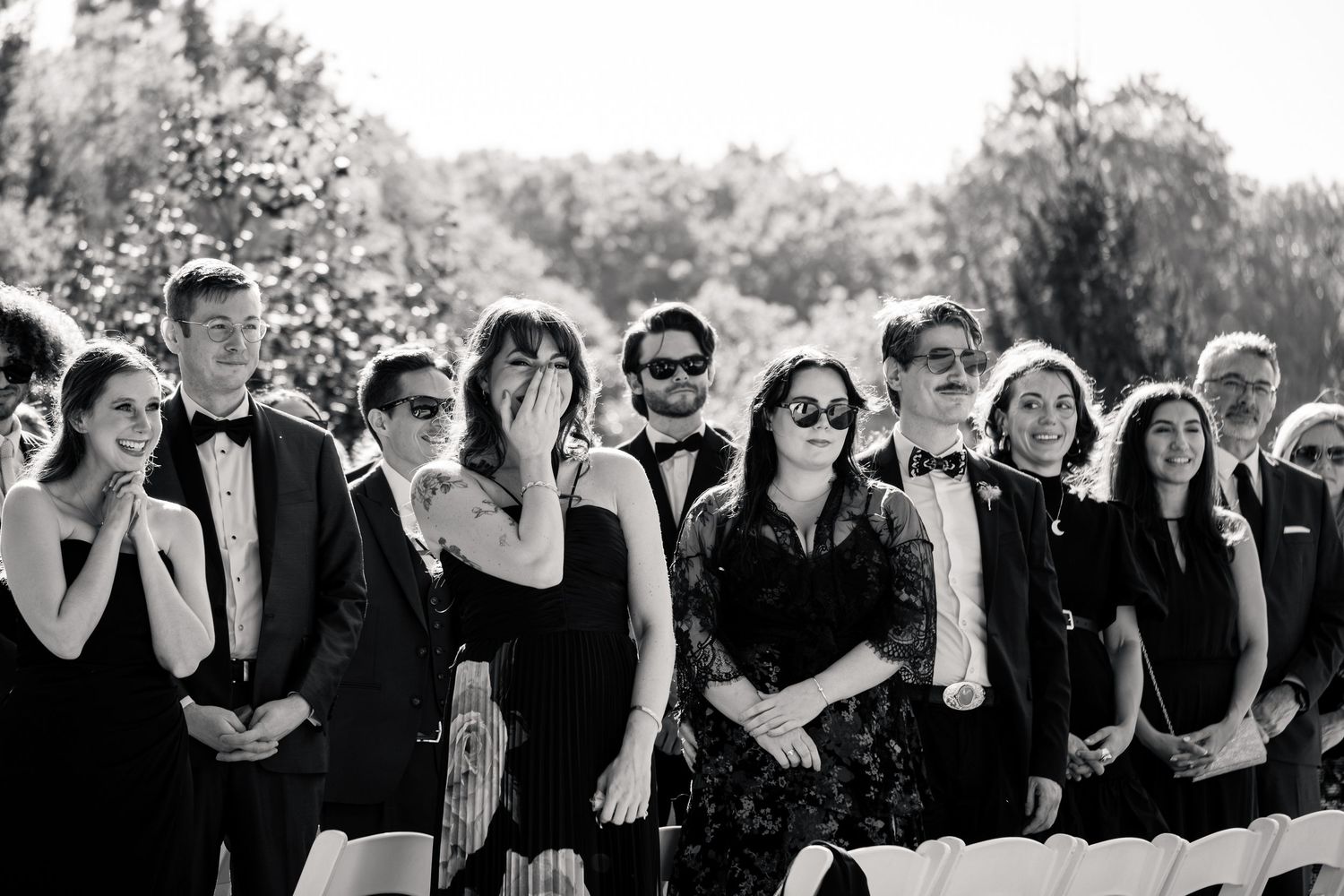 Black and white photo of emotional wedding guests watching ceremony outdoors, some crying, dressed in formal attire.