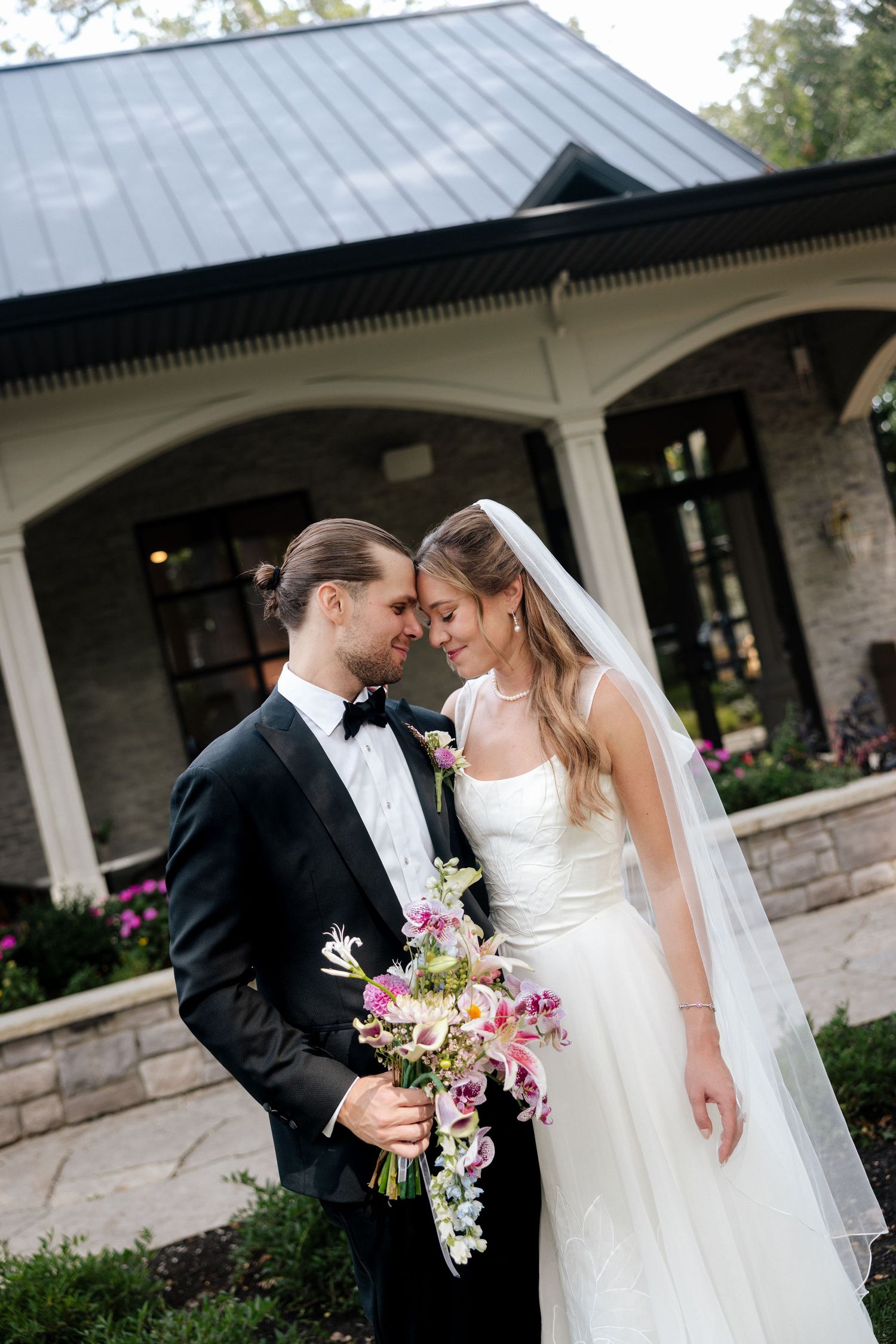 Bride and groom share a romantic moment holding a floral bouquet outside an elegant stone venue on their wedding day.