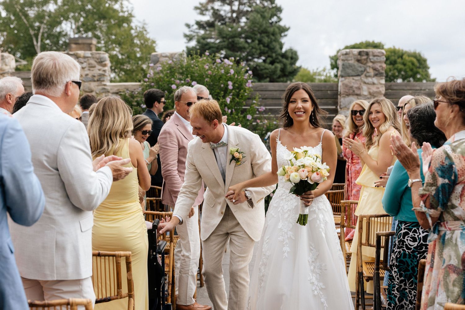 Bride and groom walk down the aisle at an outdoor wedding ceremony, guests applauding in a garden venue.