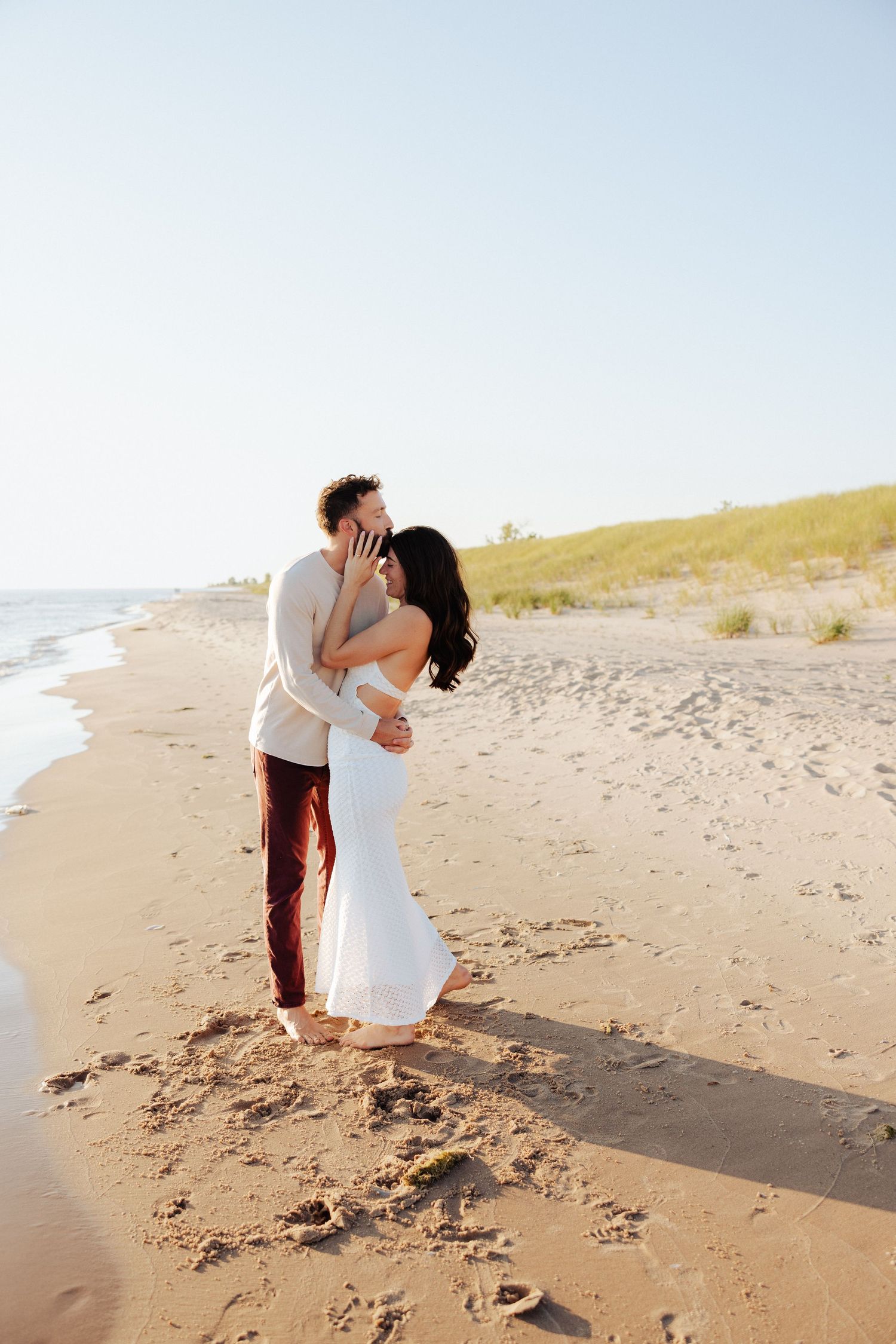 A couple shares a romantic kiss on a sandy beach, the bride in a white gown and groom in casual attire at golden hour.