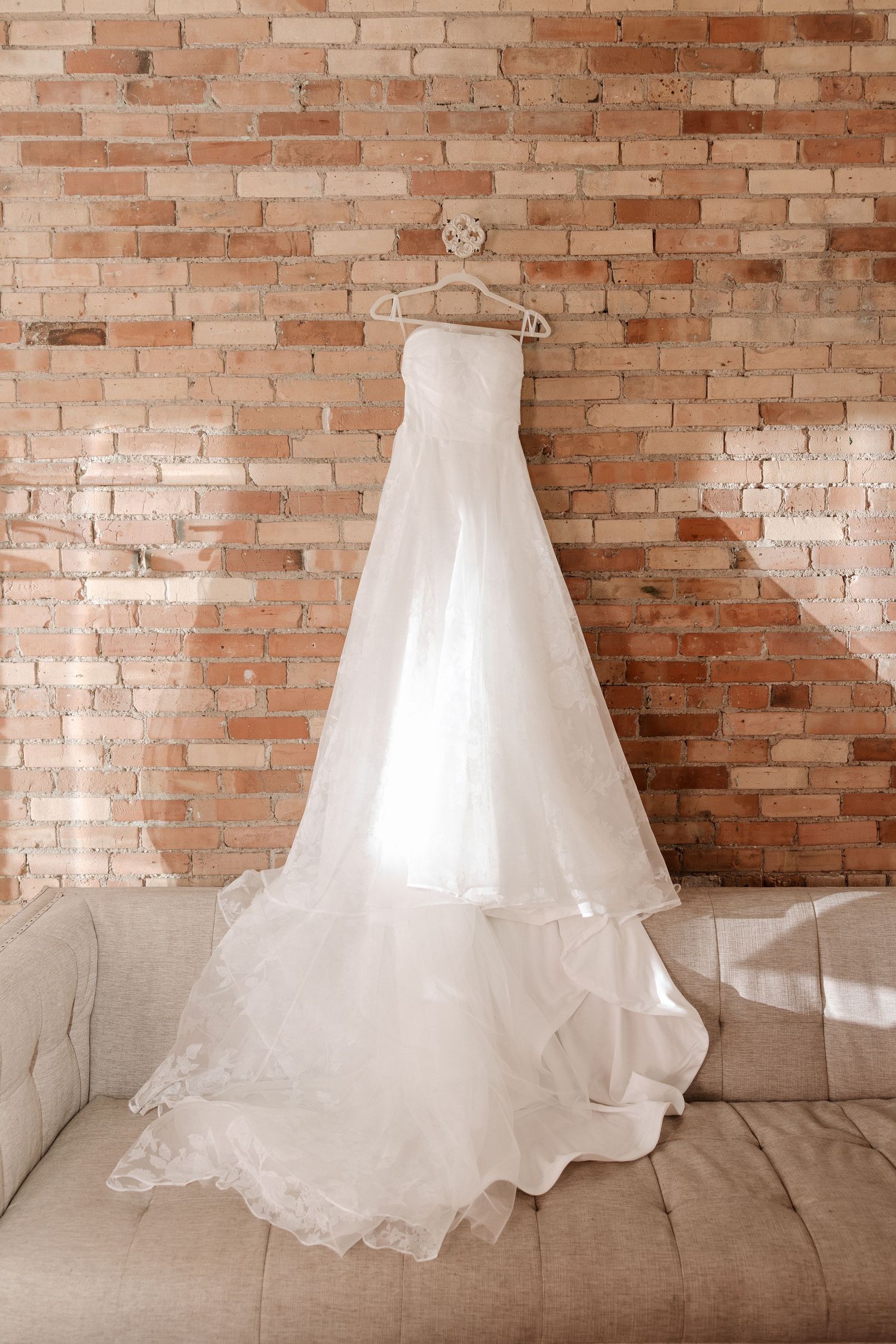 Elegant strapless white wedding gown hanging on a hanger against a rustic brick wall.
