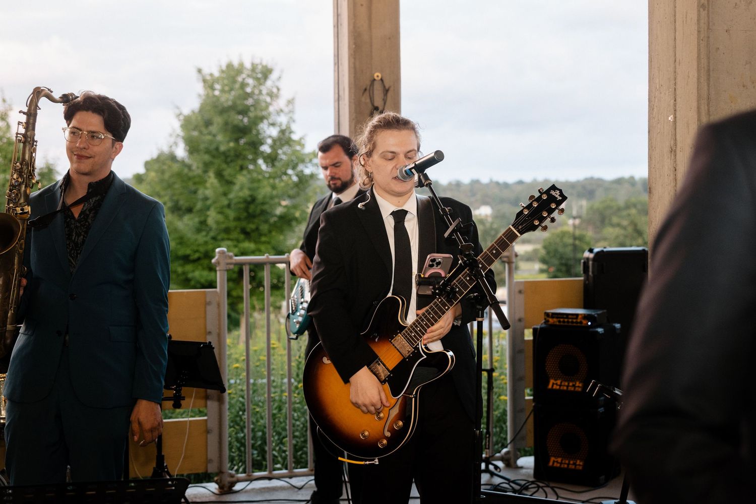 Live band performing at an outdoor event, featuring a guitarist in a black suit and saxophonist in a teal jacket.