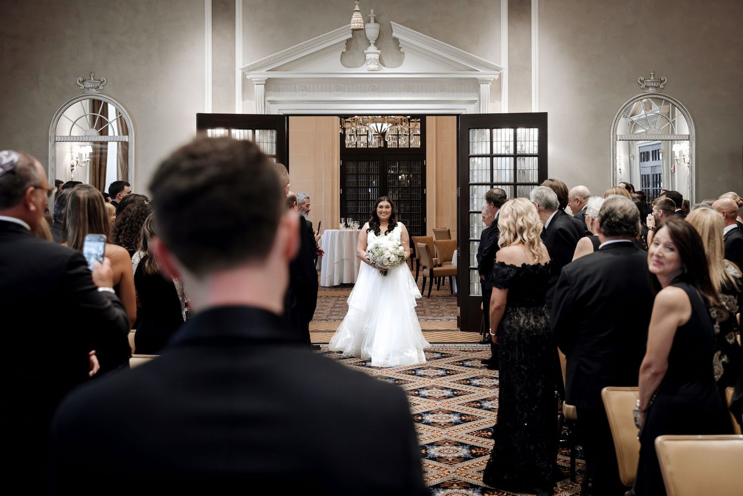 Bride walks down the aisle in a white ball gown holding a bouquet in an elegant chandelier-lit ballroom ceremony.