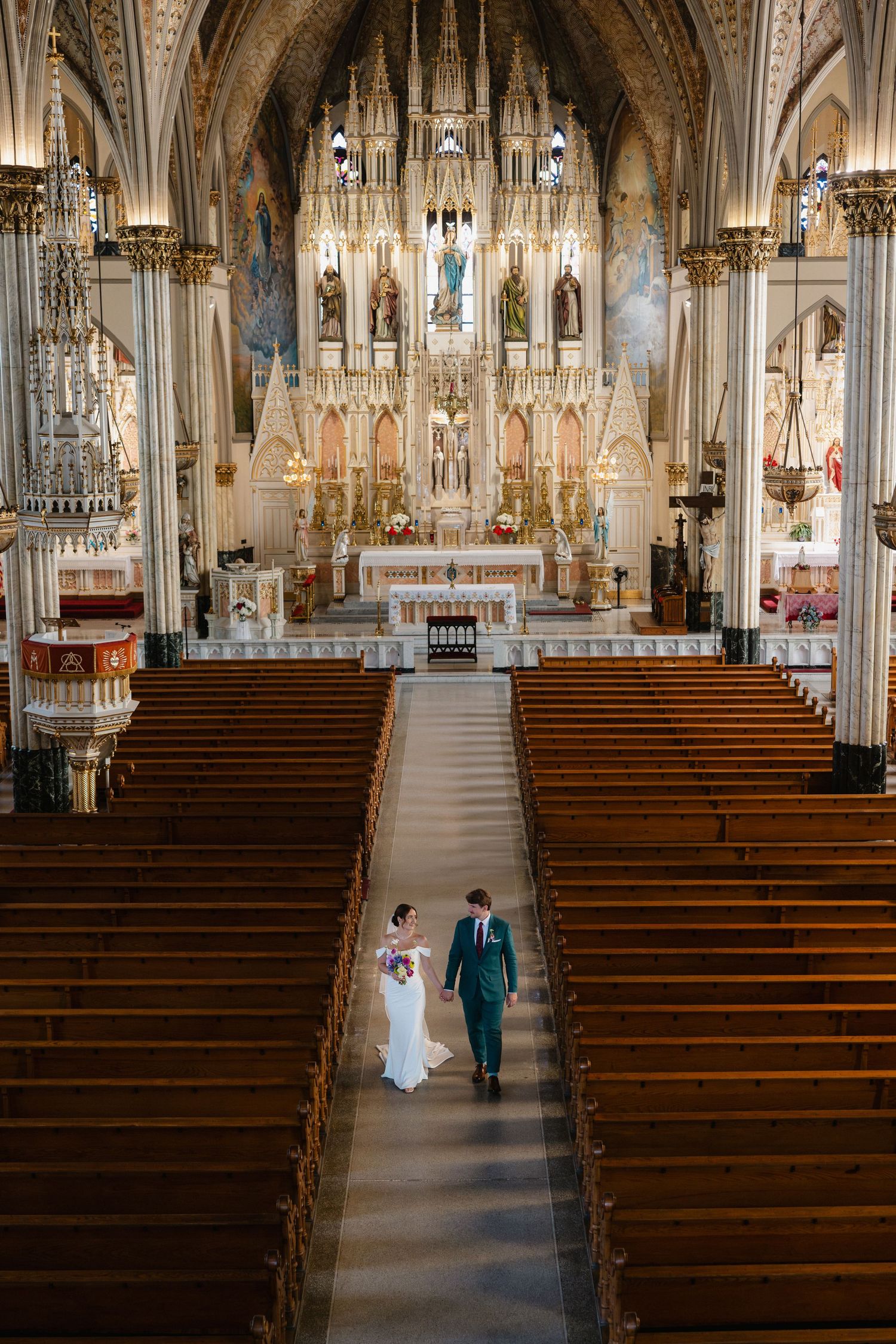 Bride and groom walk down the aisle of an ornate Gothic Catholic church with grand altar and wooden pews.