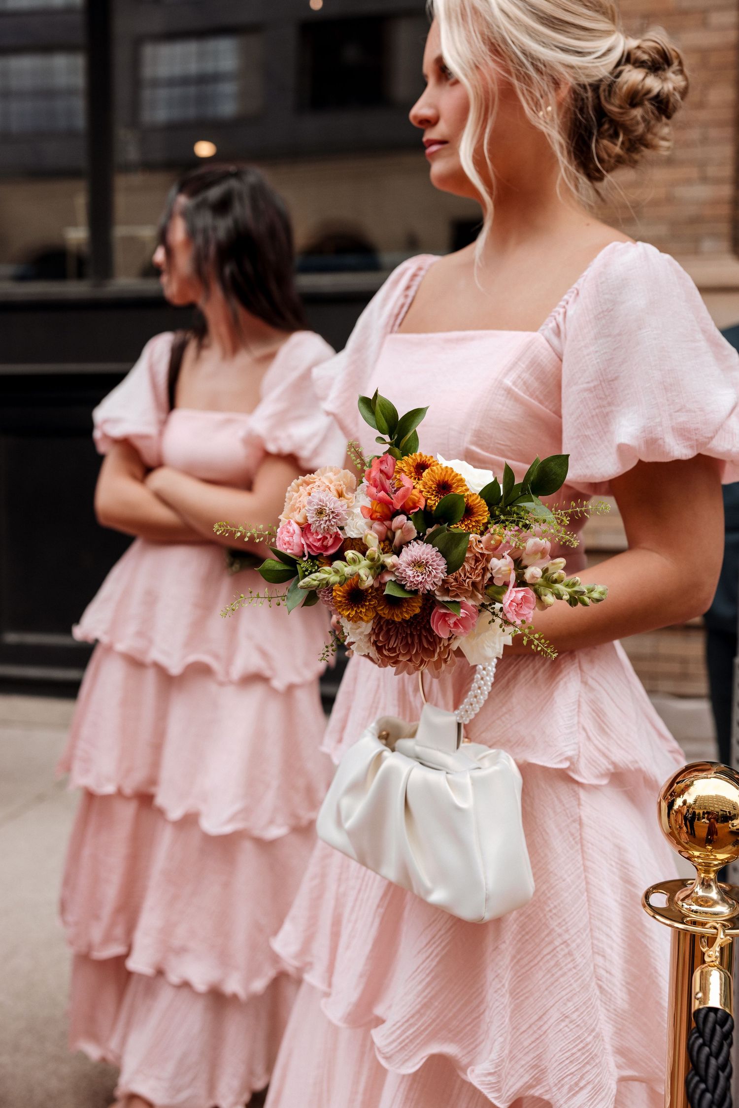 Two bridesmaids in pink ruffled dresses holding colorful floral bouquets outside venue.