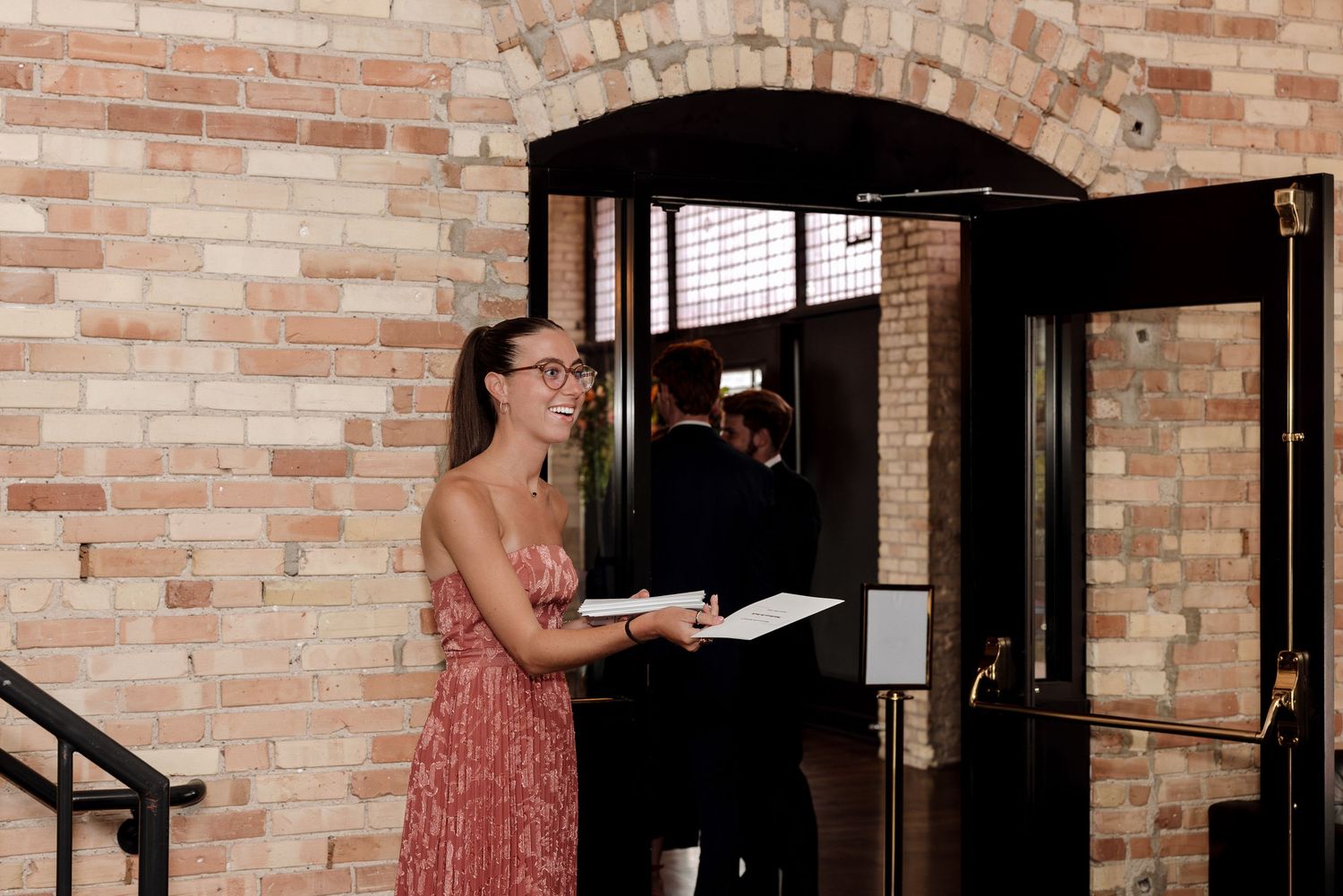 Smiling woman in pink floral dress holds a notepad while standing at an elegant brick archway entrance.
