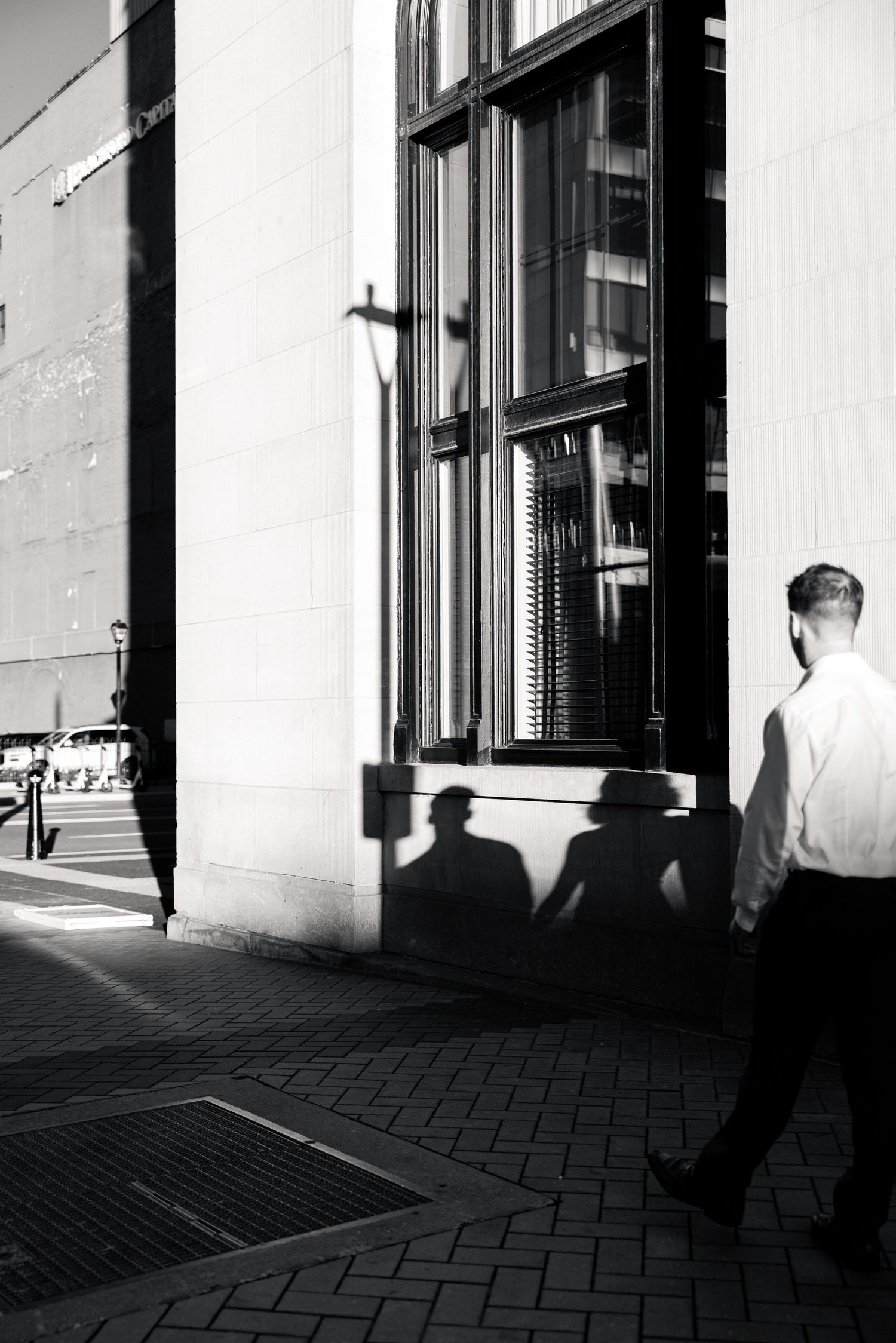 Black and white street photo of a man walking past a building, with dramatic shadows cast on the wall beside him.