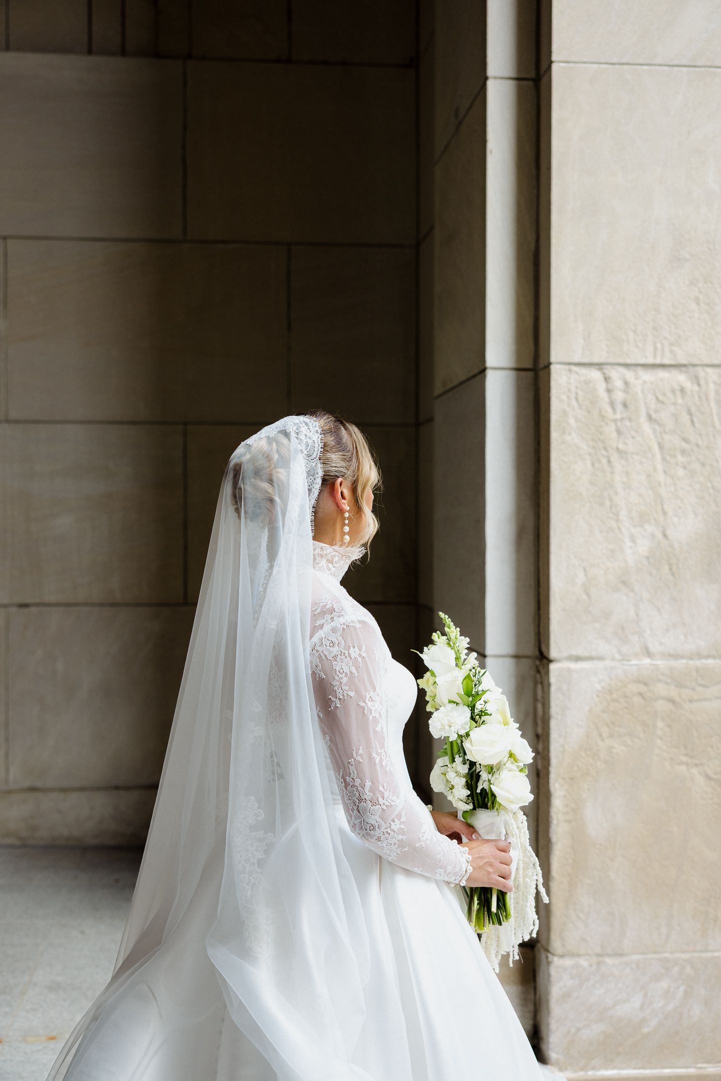 Bride in lace long-sleeve wedding gown and veil holds white bouquet, standing by stone columns in elegant venue.