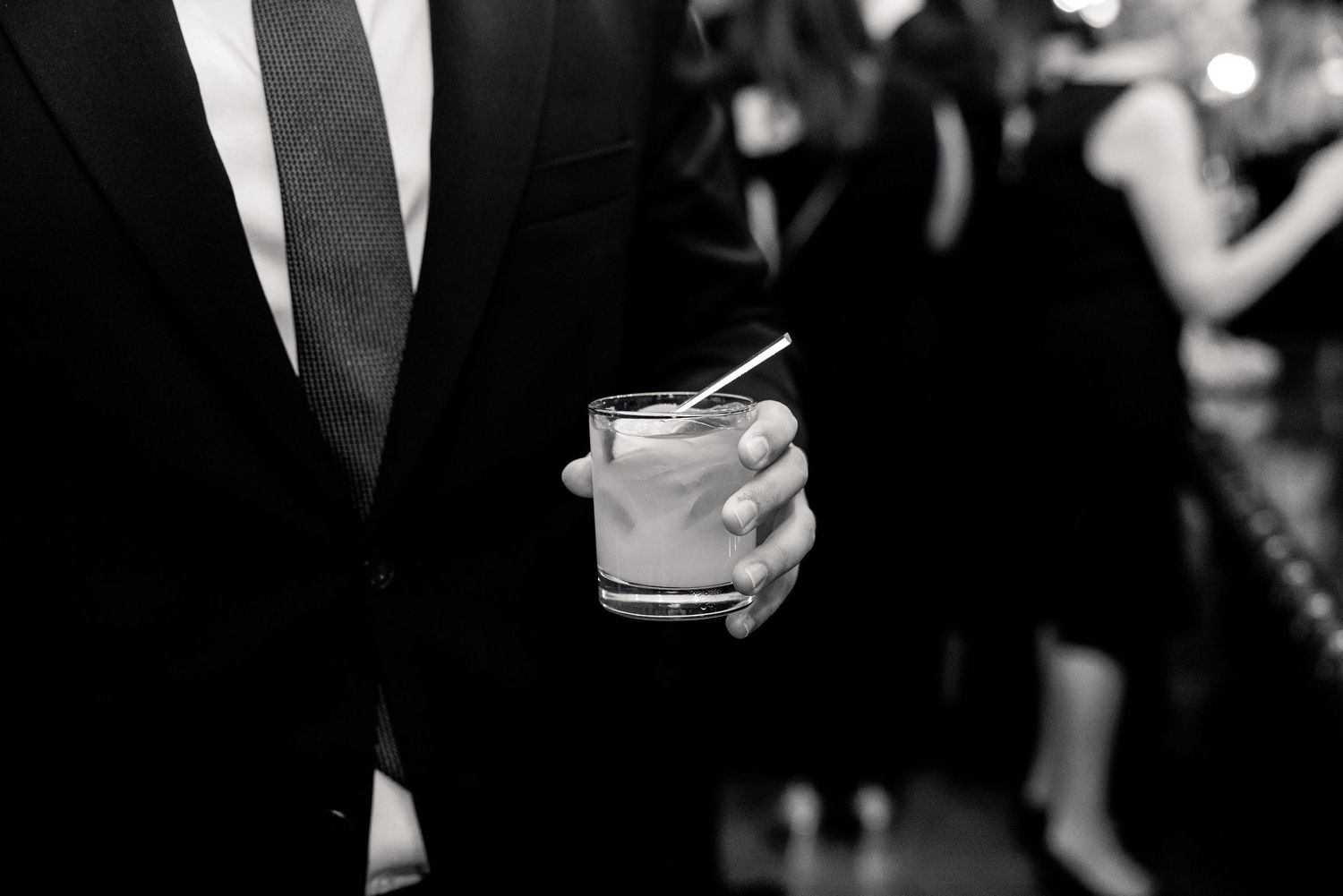 Close-up of a man in a black tuxedo holding a cocktail glass at a formal wedding reception.
