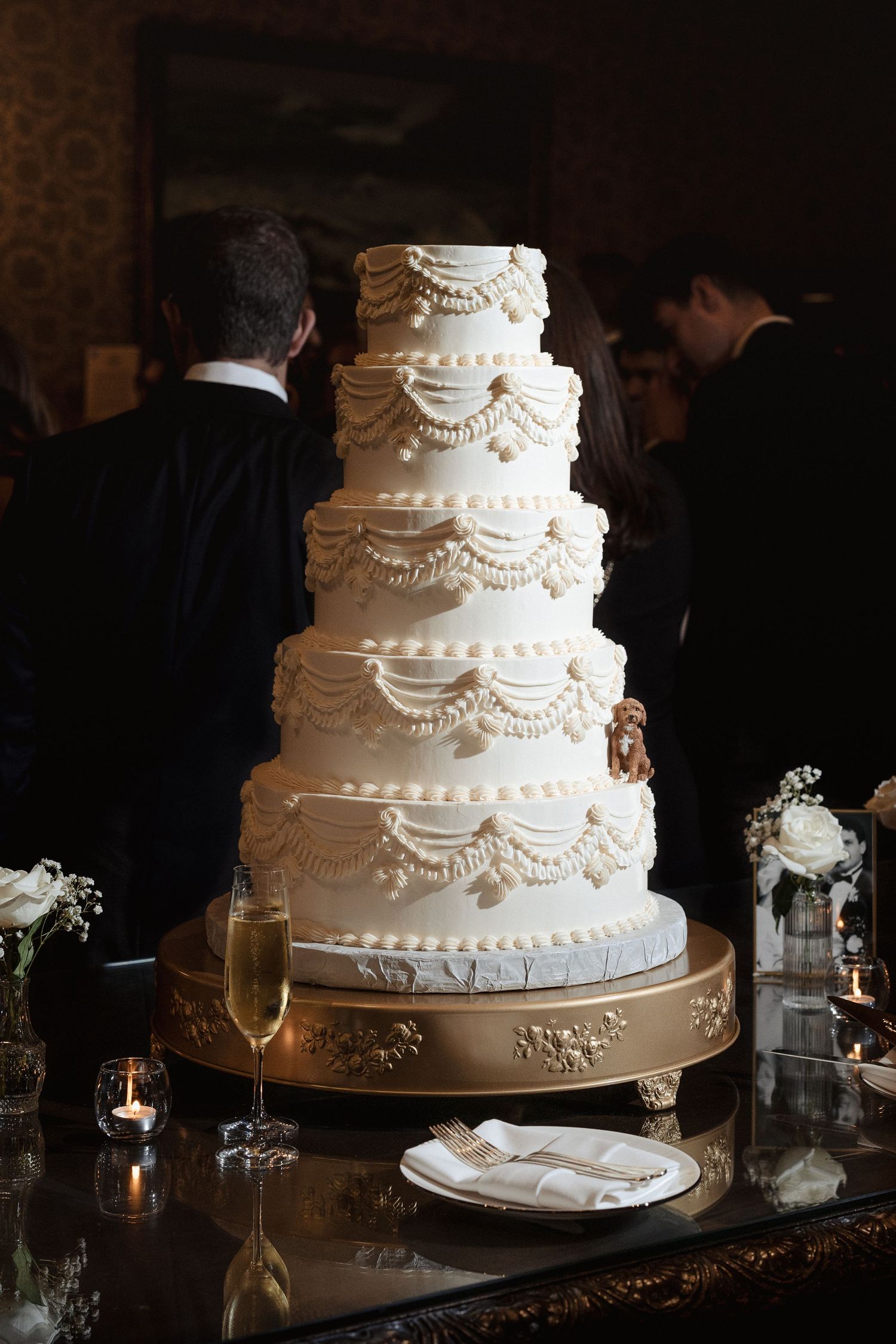 Stunning six-tier white wedding cake with pearl swag details on a gold stand at a black tie reception.