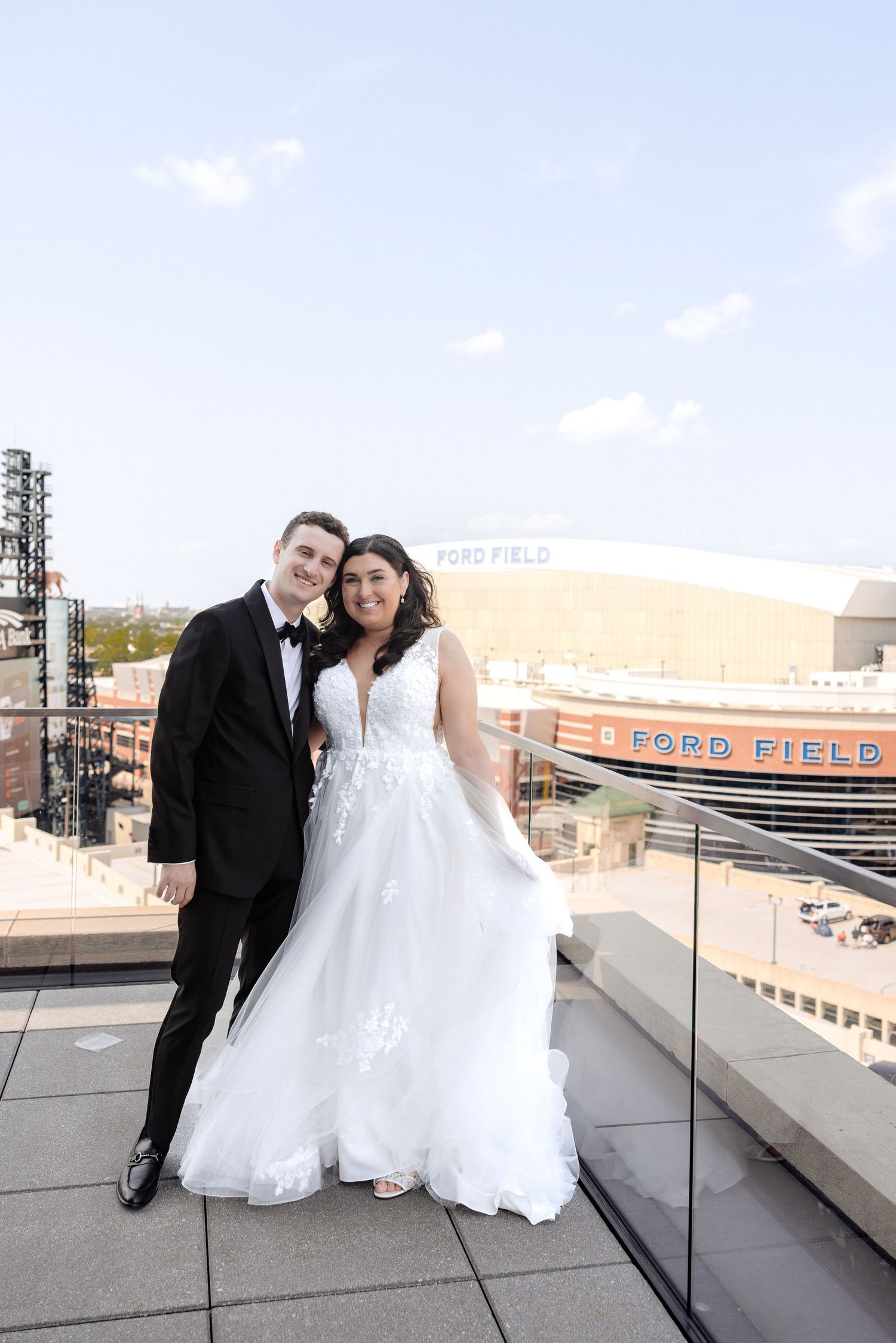 Happy newlyweds embracing on a rooftop terrace with Ford Field stadium visible behind them.
