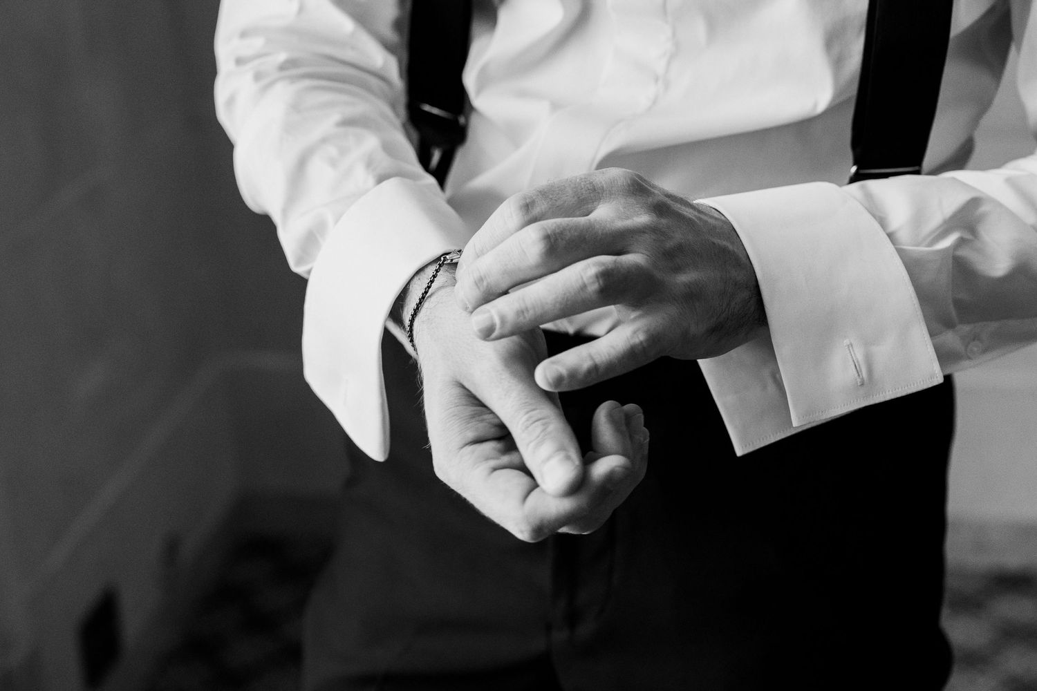 Black and white close-up of groom adjusting cufflinks on white dress shirt with black suspenders.