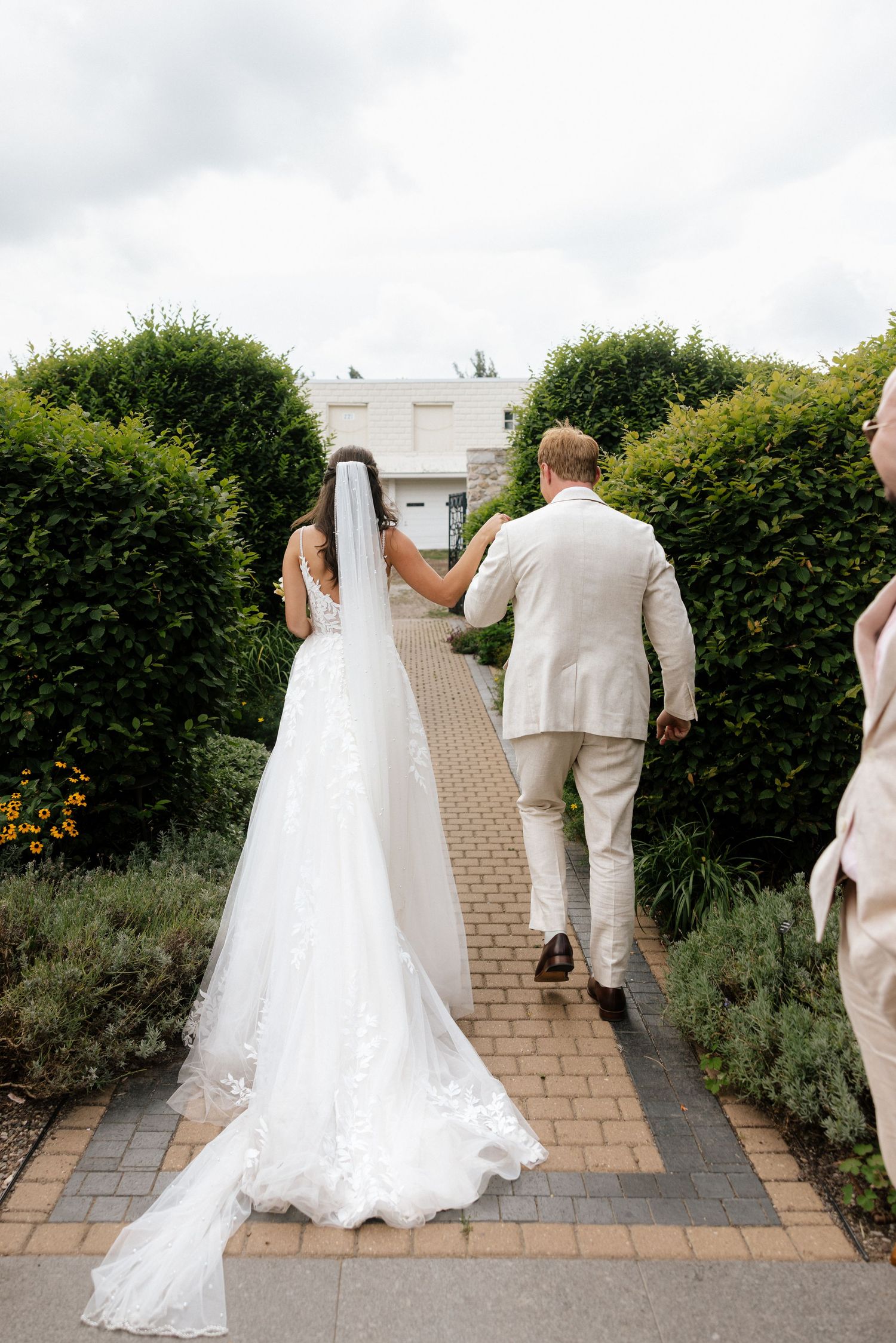 Bride and groom walking hand-in-hand down a brick path lined with tall green hedges.