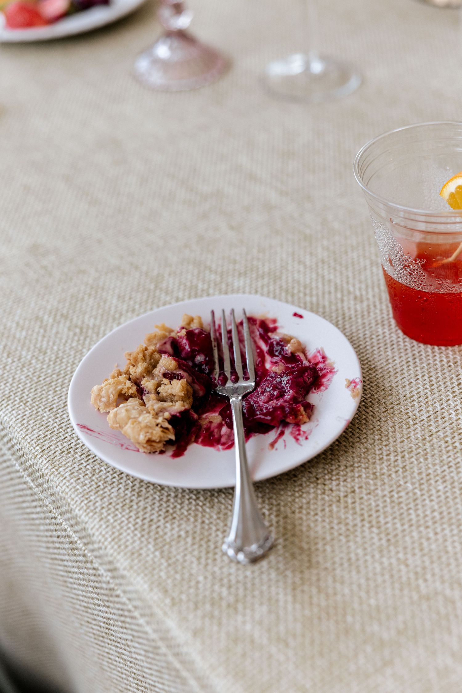 Partially eaten berry crumble dessert on white plate with fork beside a cocktail glass at an event.