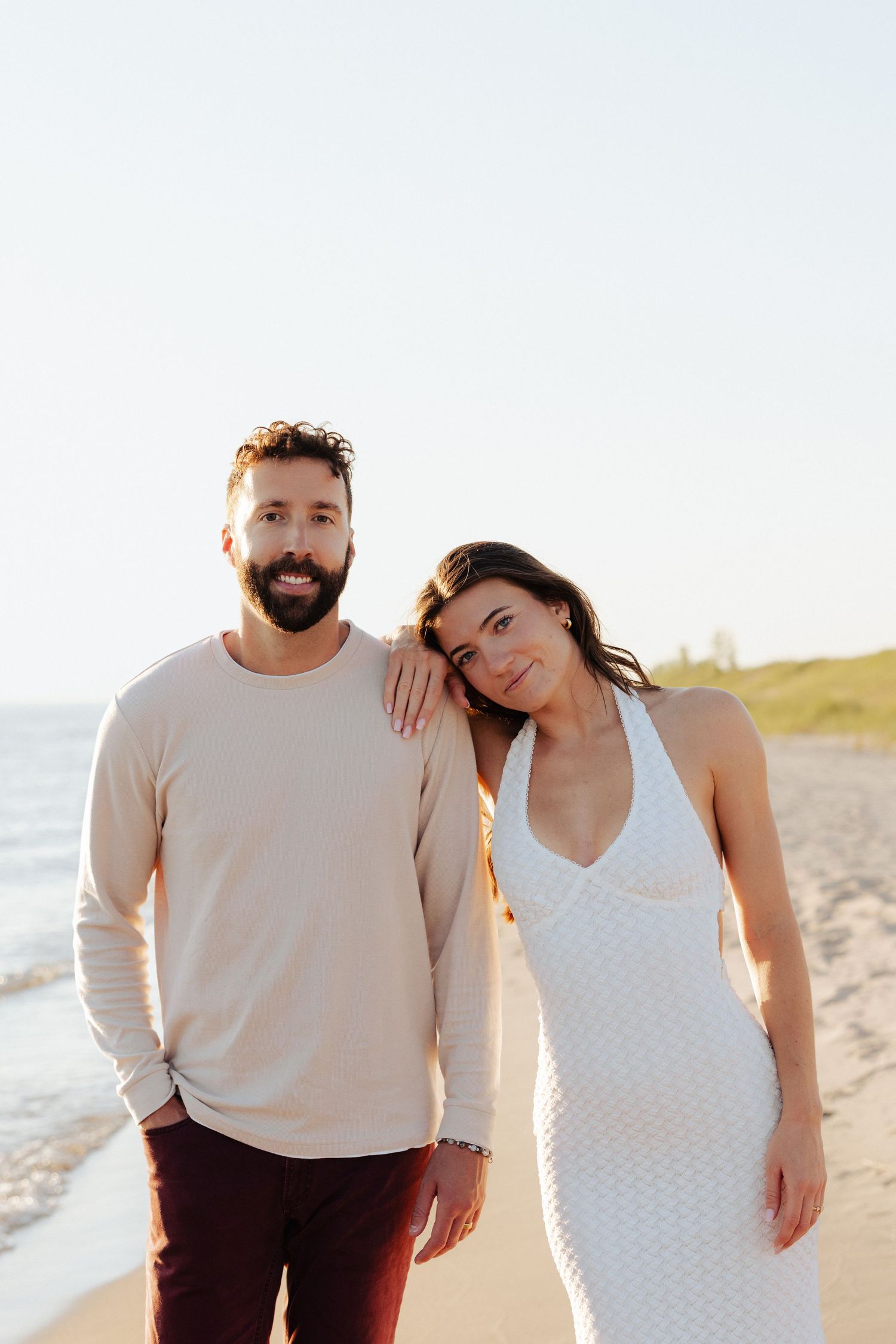 A smiling couple poses on a sunny beach, man in beige top and woman in white dress leaning on his shoulder.