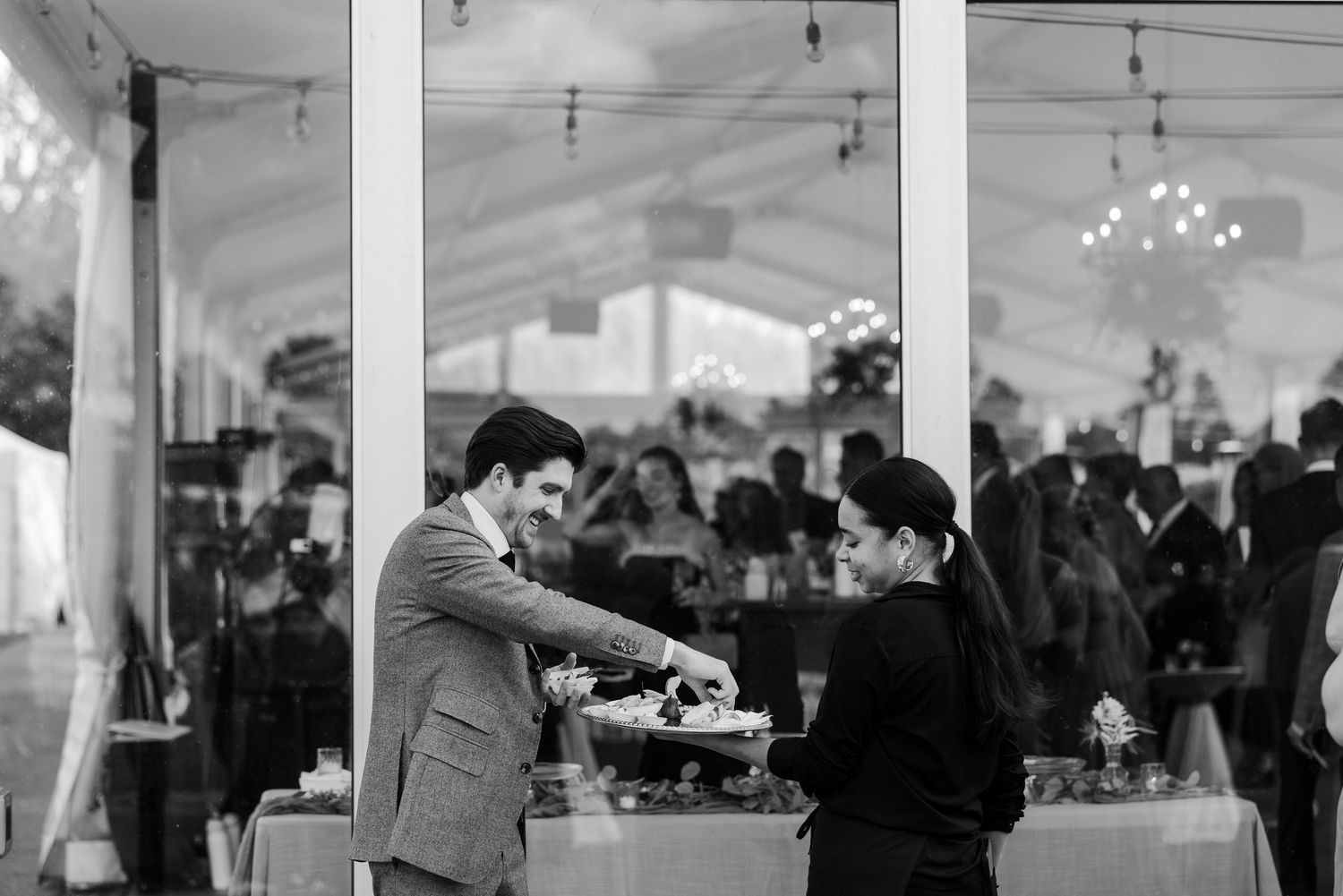 Black and white photo of a man in a suit being served at an elegant outdoor wedding reception tent.