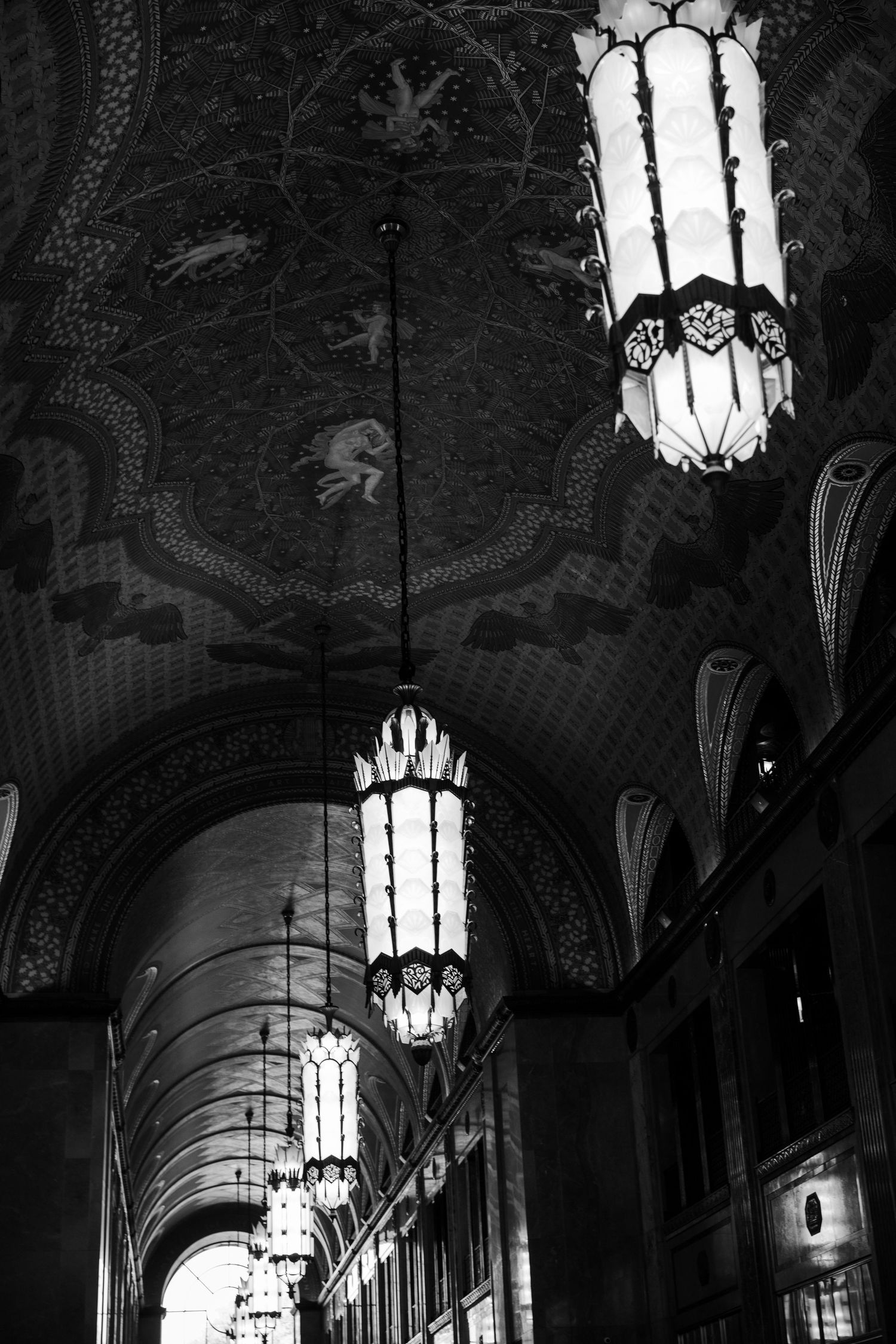 Black and white photo of ornate ceiling with hanging lanterns in an elegant arched corridor.