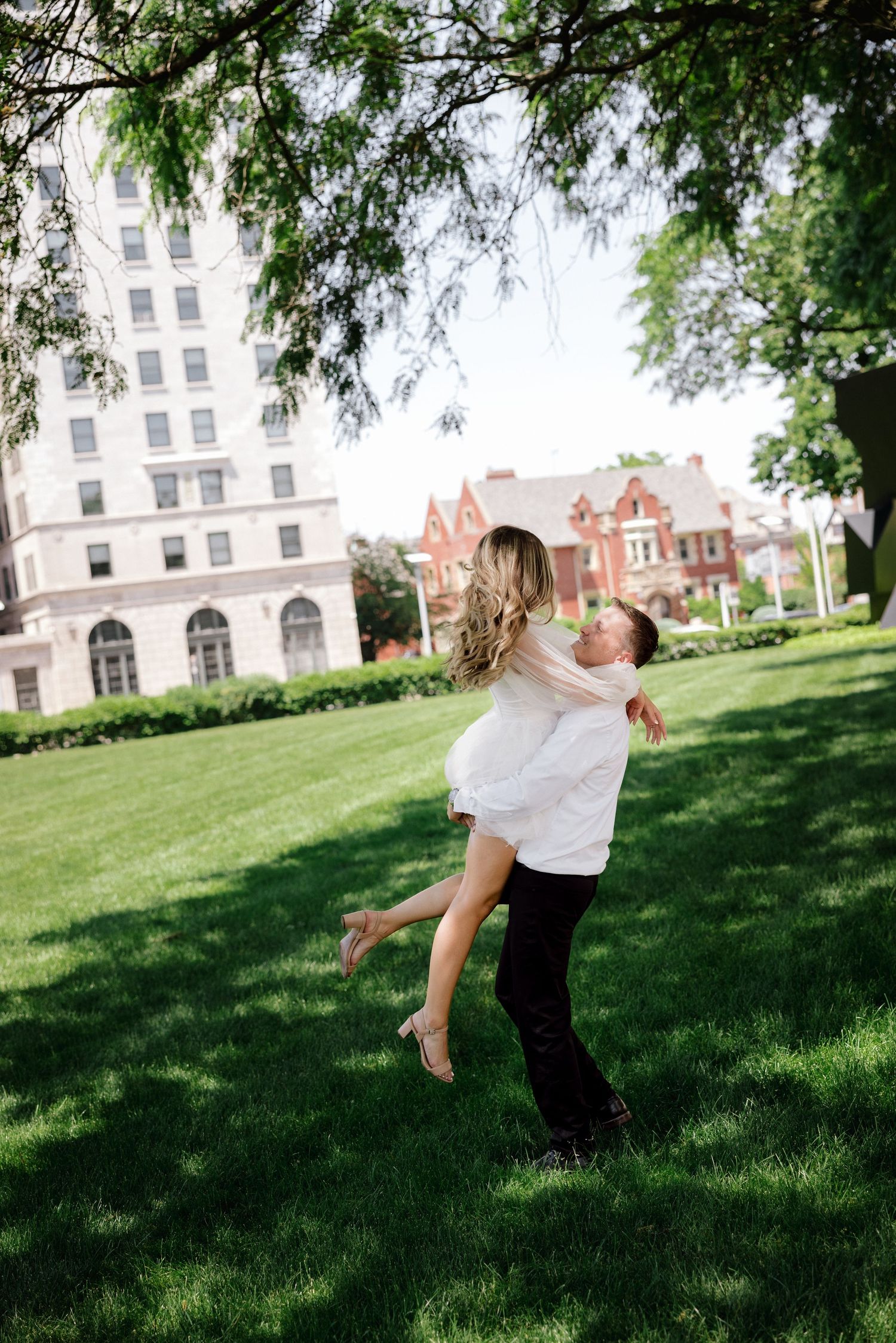 Man lifting and spinning woman in white dress on lush green lawn with historic buildings behind.